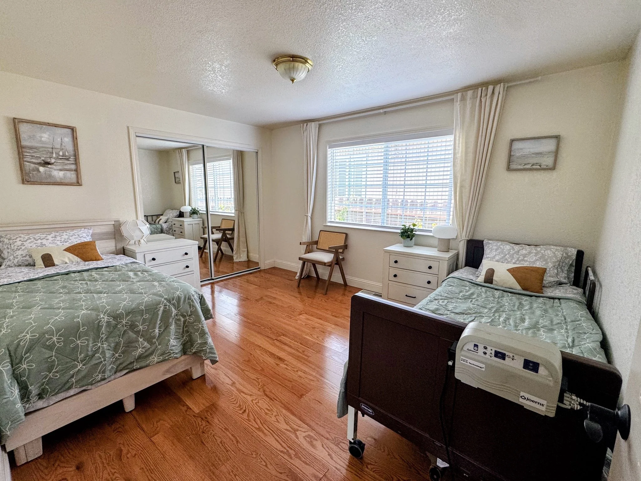 A bedroom with two beds, white furniture, a large window with blinds, and a mirror closet door. The room has wooden floors, wall art, and a small fan on one of the beds.