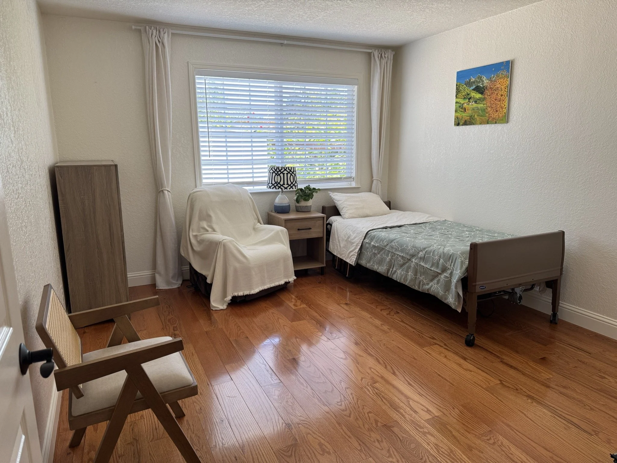 A small bedroom with a wood floor, a bed with a comforter, a nightstand with a potted plant and a lamp, a chair with a blanket, a wooden wardrobe, a framed landscape painting on the wall, and a large window with blinds and curtains.