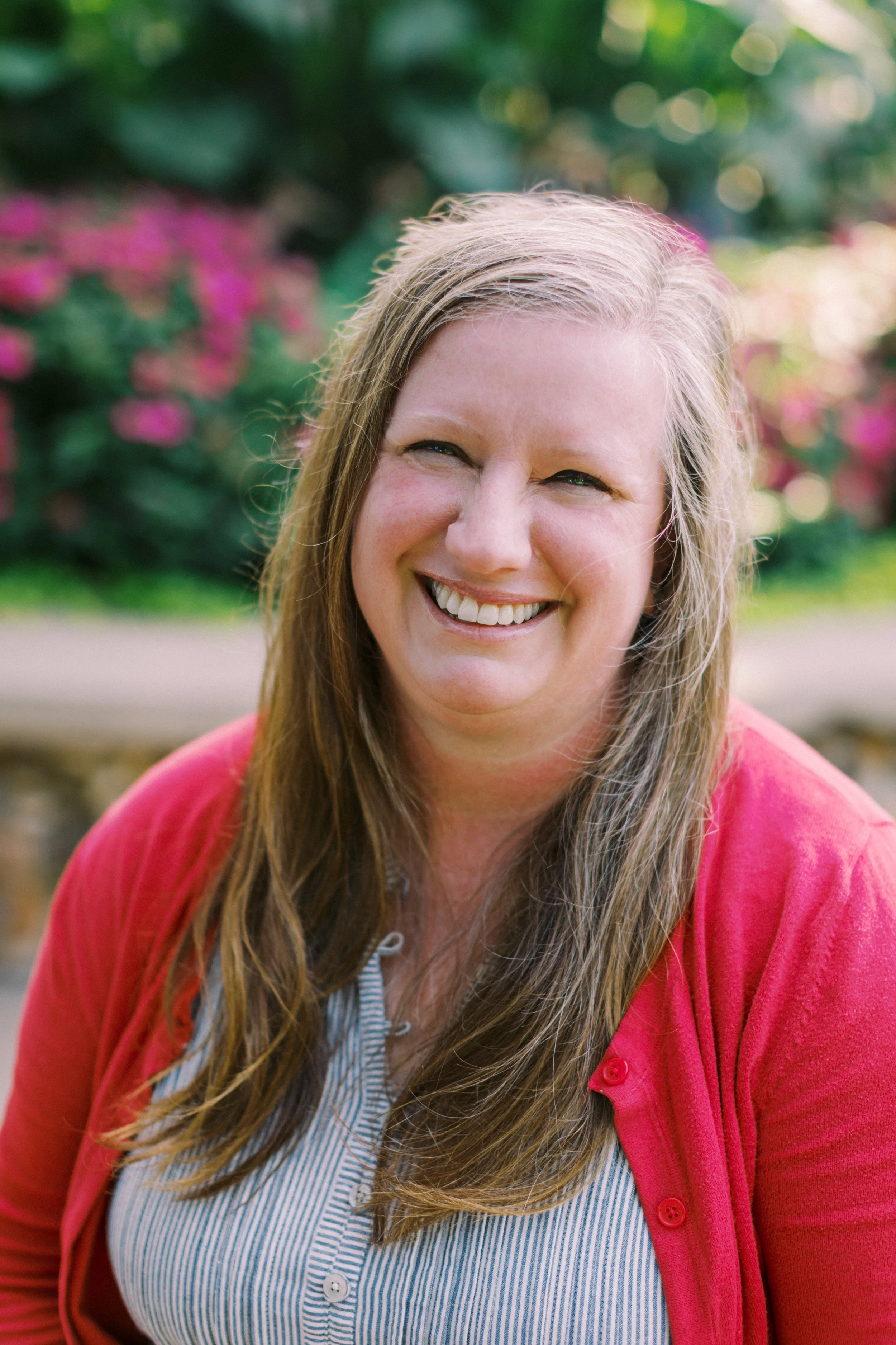 Smiling woman with long hair wearing a red cardigan and striped shirt, outdoors with blurred flowers and greenery in the background.