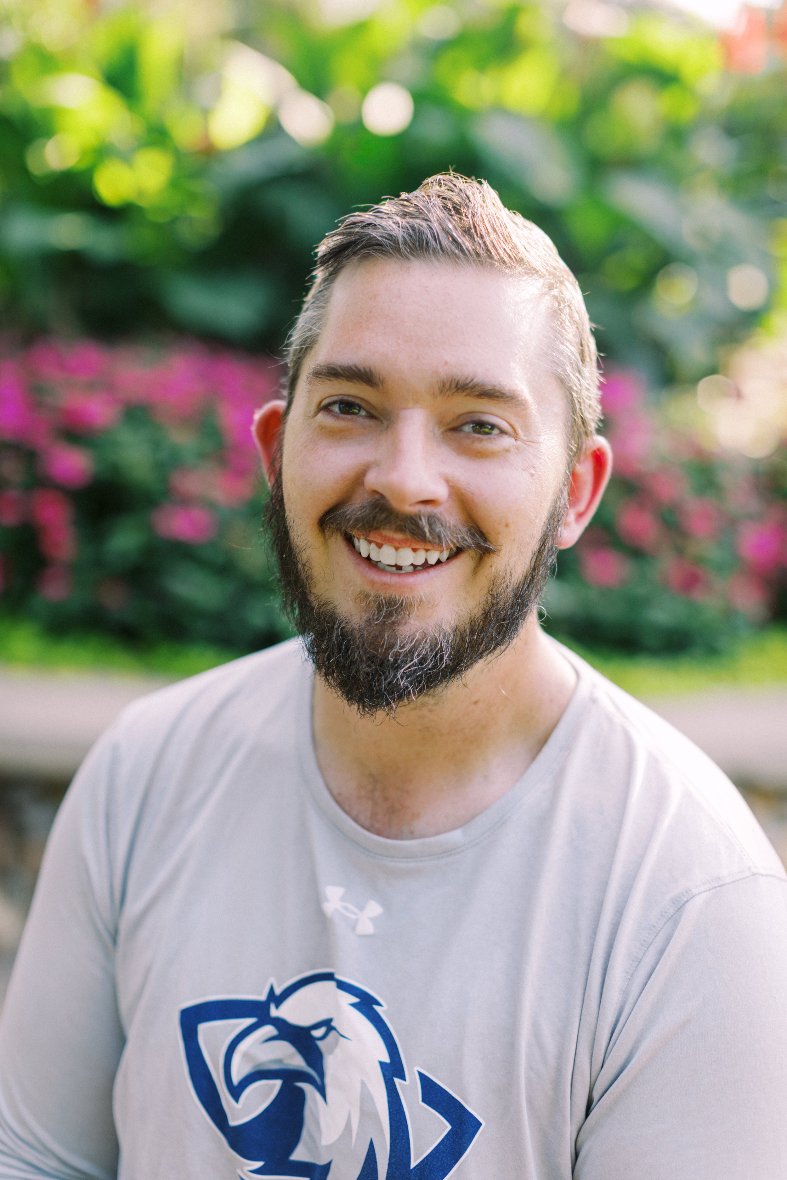 A smiling man with a beard and mustache wearing a gray athletic shirt with a bird logo, outdoors with greenery and pink flowers in the background.