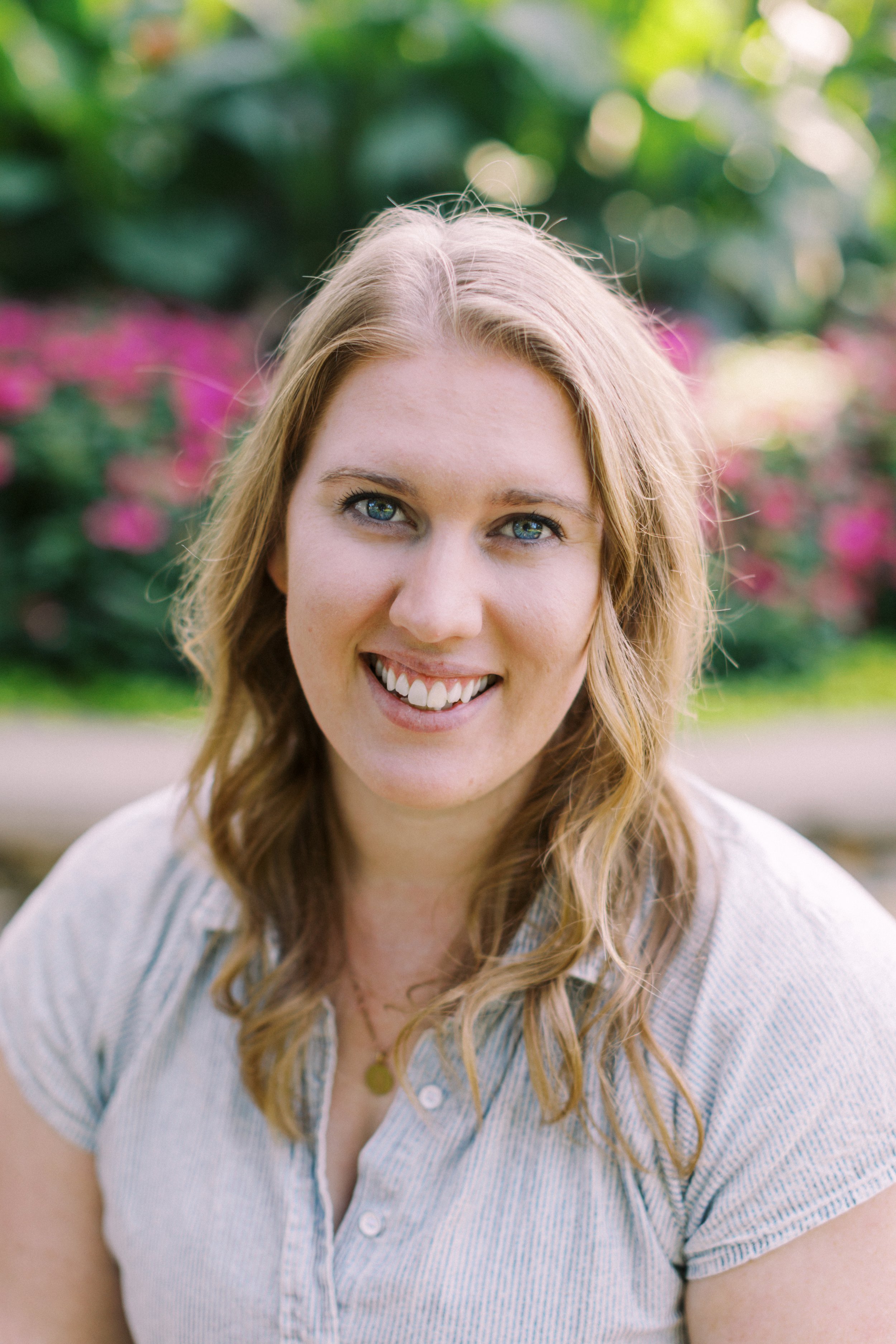 A woman with blonde hair and blue eyes smiling outdoors with blurred pink flowers and green foliage in the background.