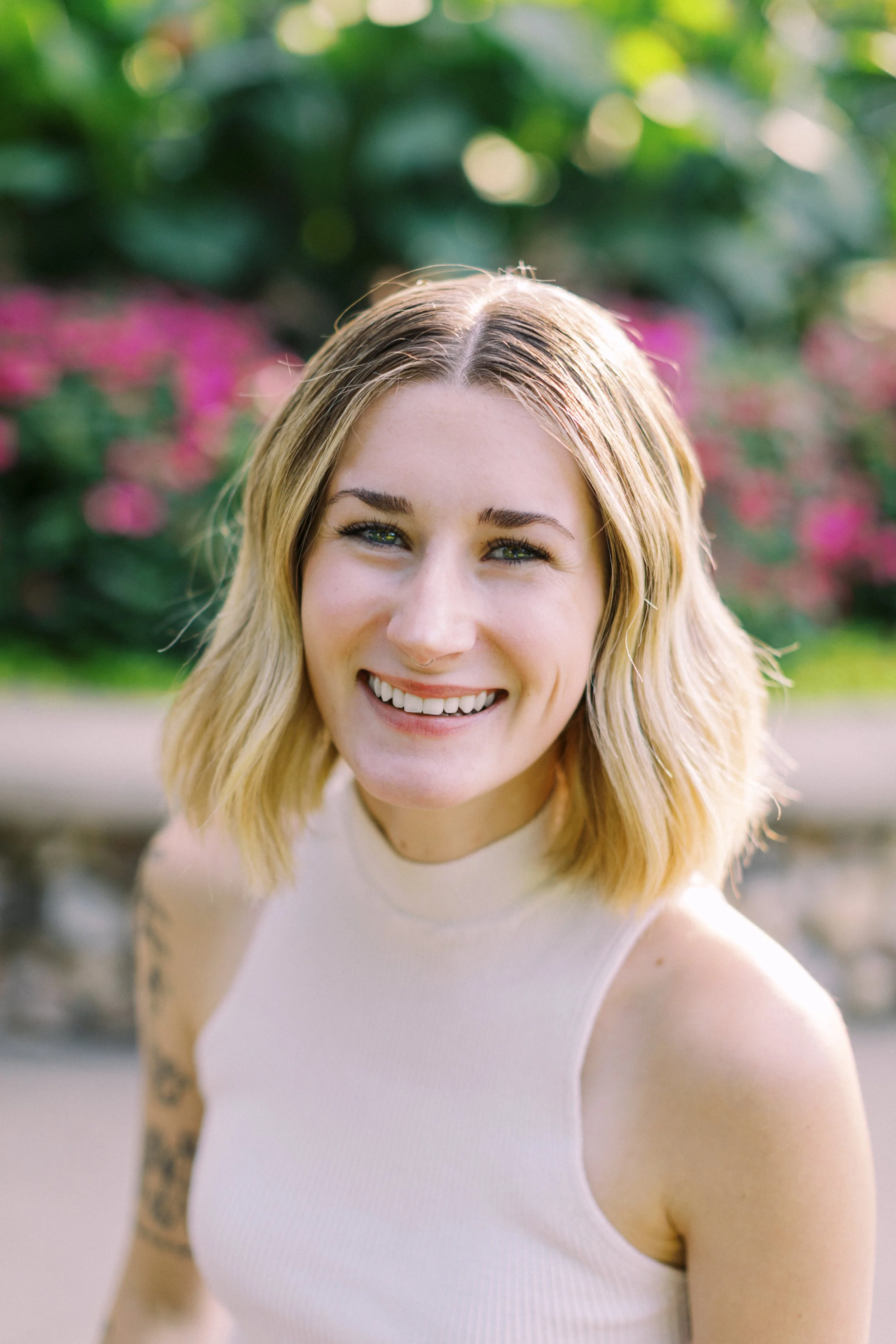 A smiling woman with blonde hair standing outdoors with greenery and pink flowers in the background.
