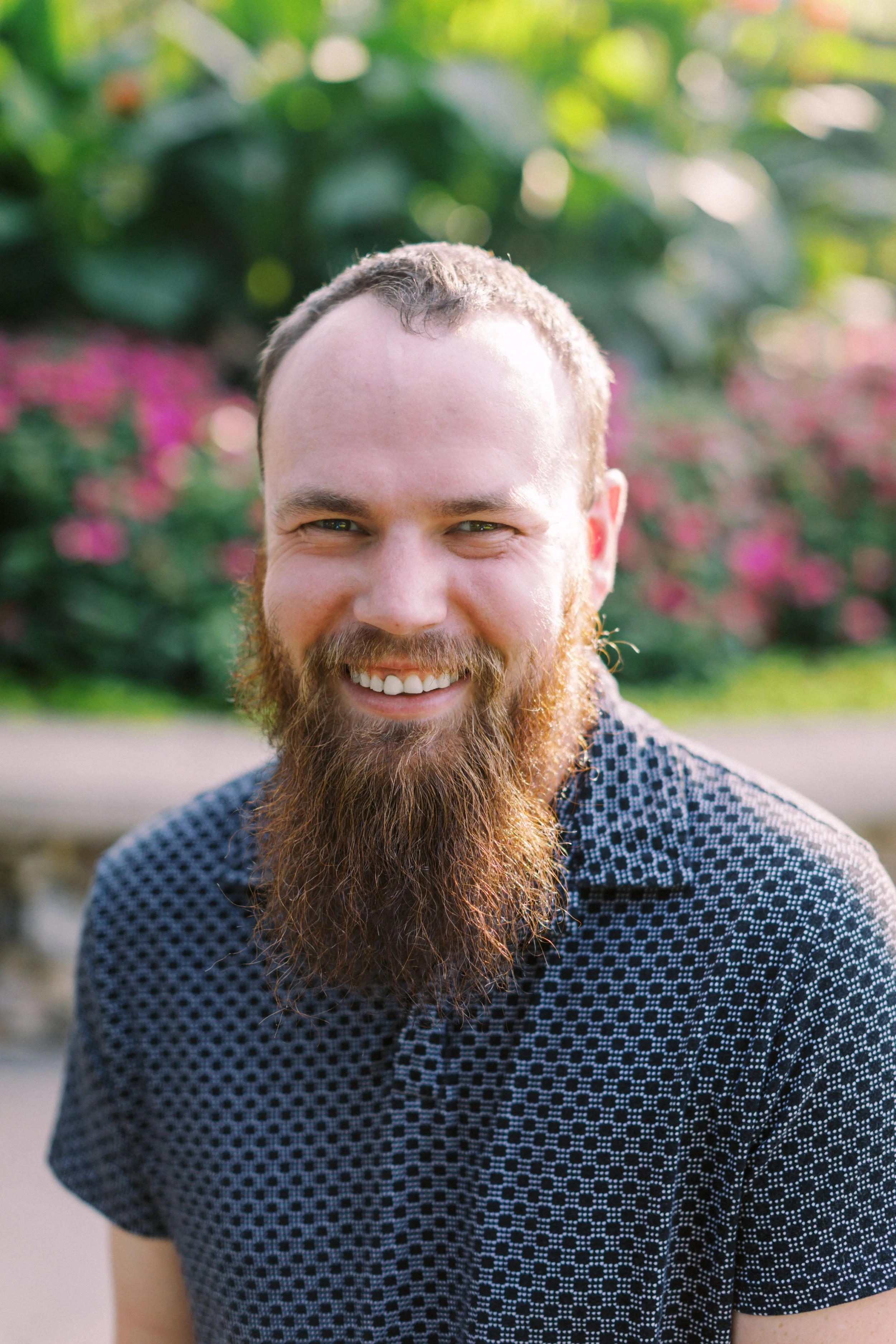 A smiling man with a full beard and short hair, wearing a patterned short-sleeve shirt, standing outdoors in front of blurred greenery and pink flowers.