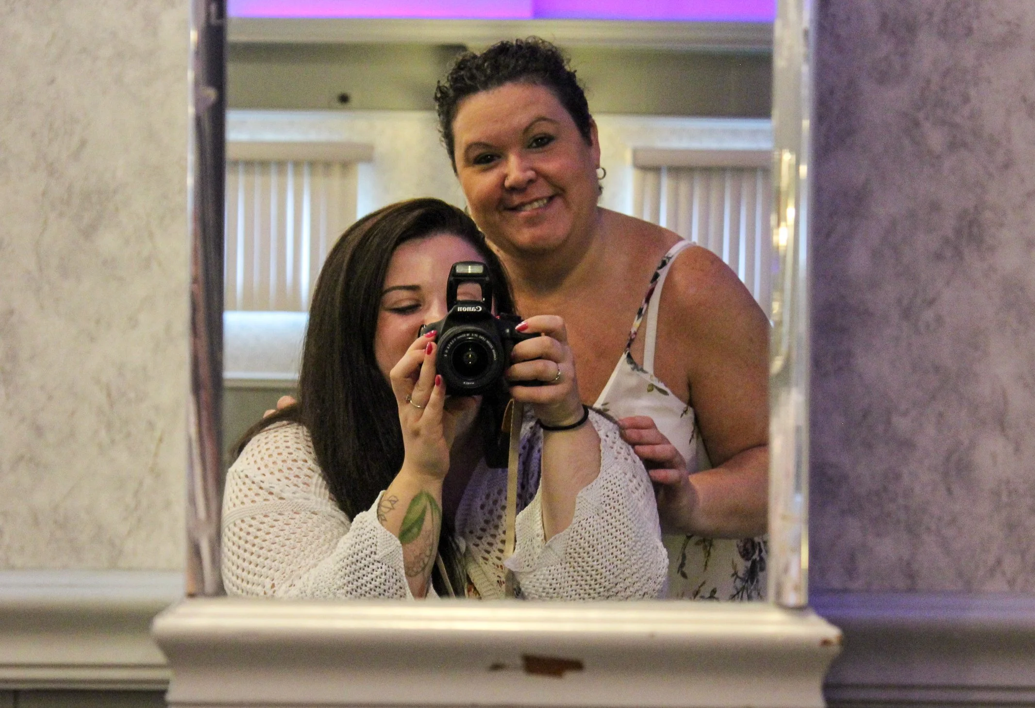 Two women taking a selfie in a mirror. The woman in front, with dark hair, is holding a camera, and the woman behind with short curly hair is smiling.
