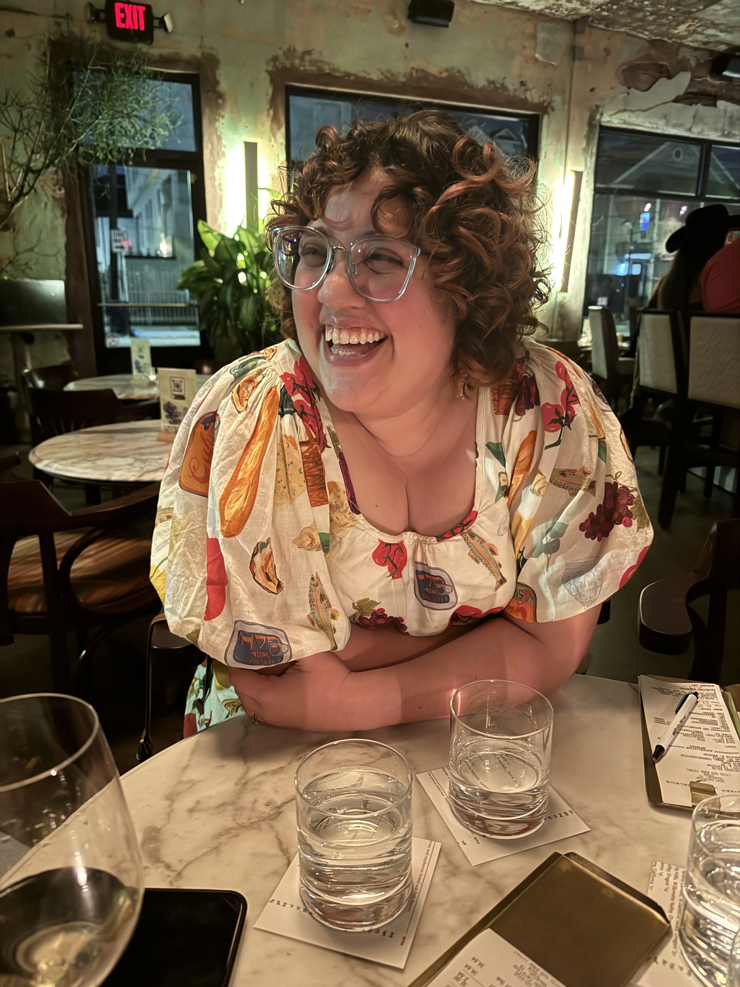 A woman with curly brown hair and big glasses, wearing a colorful patterned blouse, sitting at a table laughing in a restaurant with a rustic interior and large windows.
