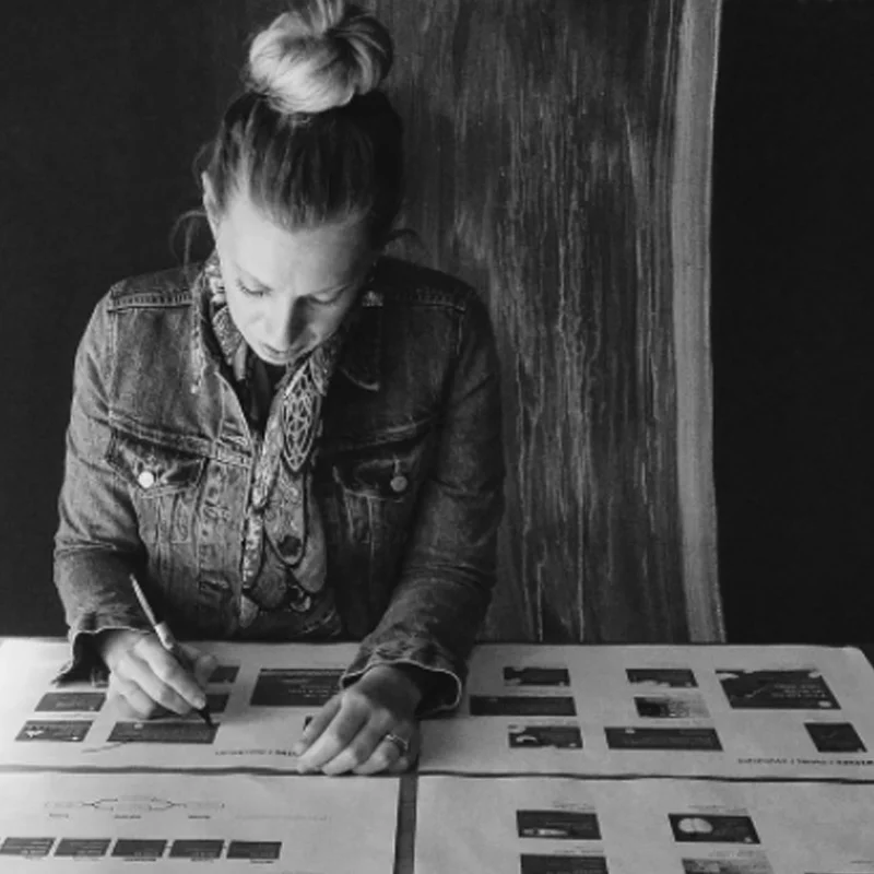 Woman reviewing design layouts on paper, taking notes, wearing a denim jacket, black and white photo.