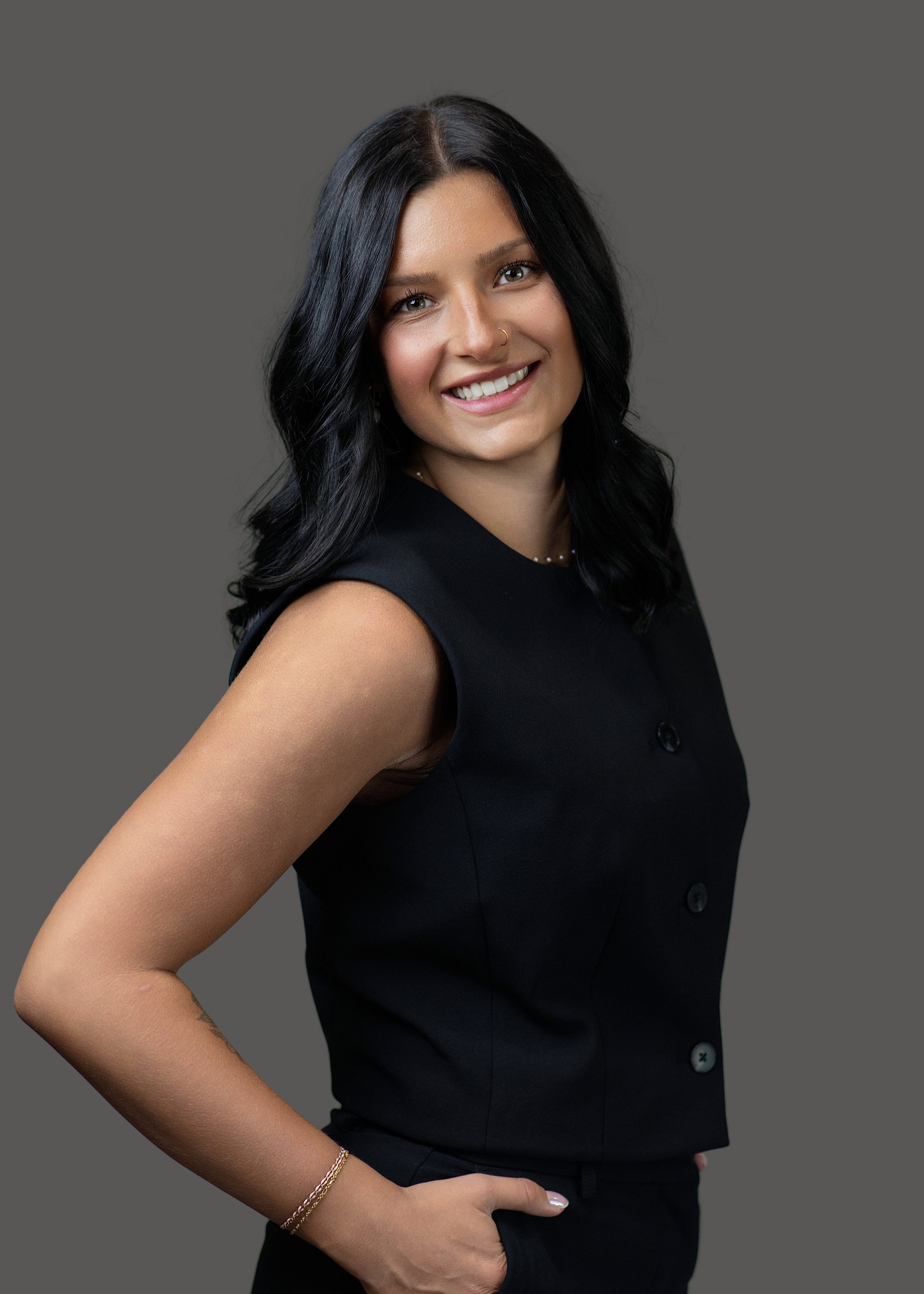 Close-up of a smiling woman with long black hair, wearing a black sleeveless button-up top, a gold bracelet, and a nose ring, standing against a plain gray background.