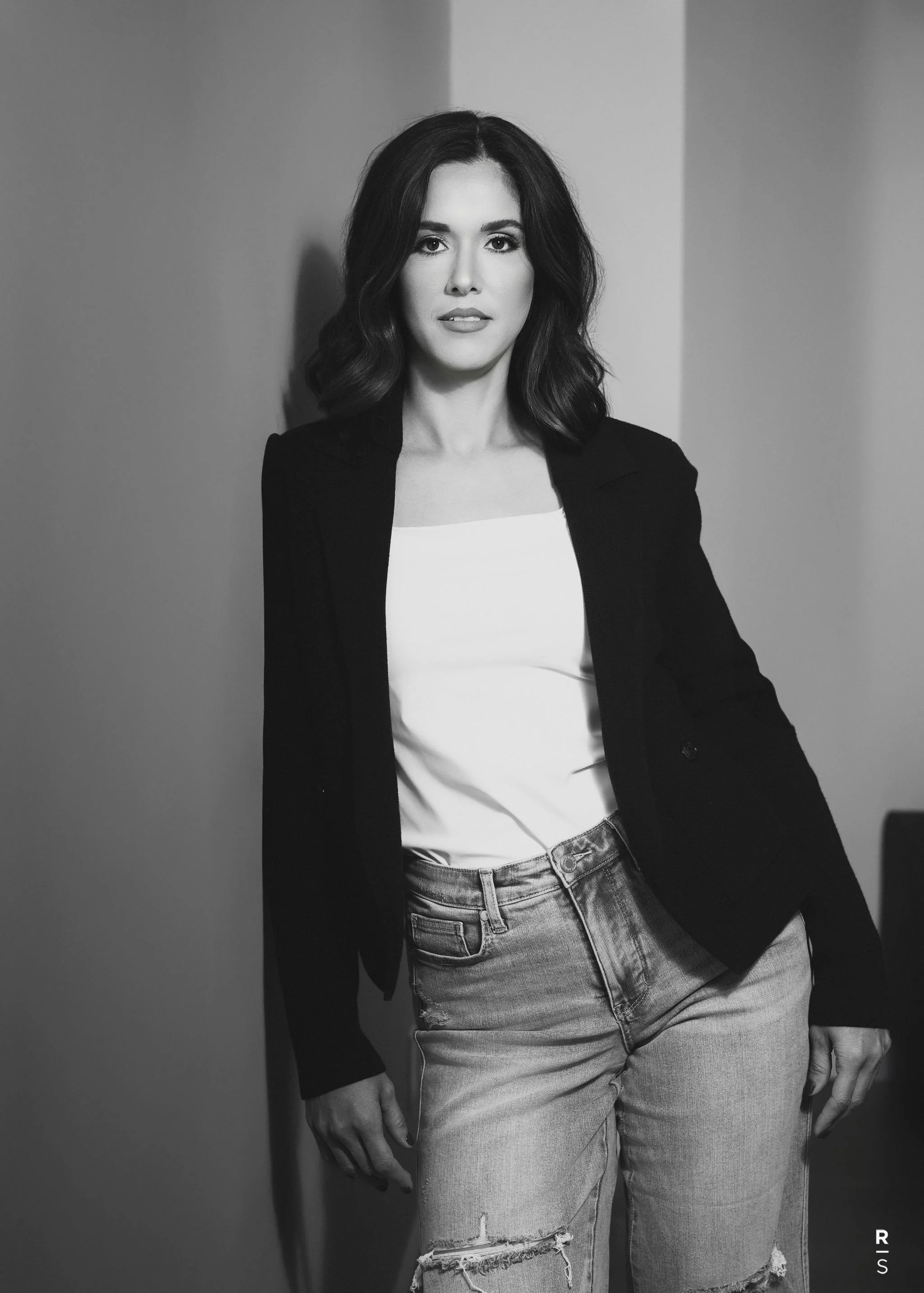 Black and white photo of a woman with wavy dark hair, wearing a blazer and white shirt, standing against a plain wall with one arm at her side, and the other hand partly visible.
