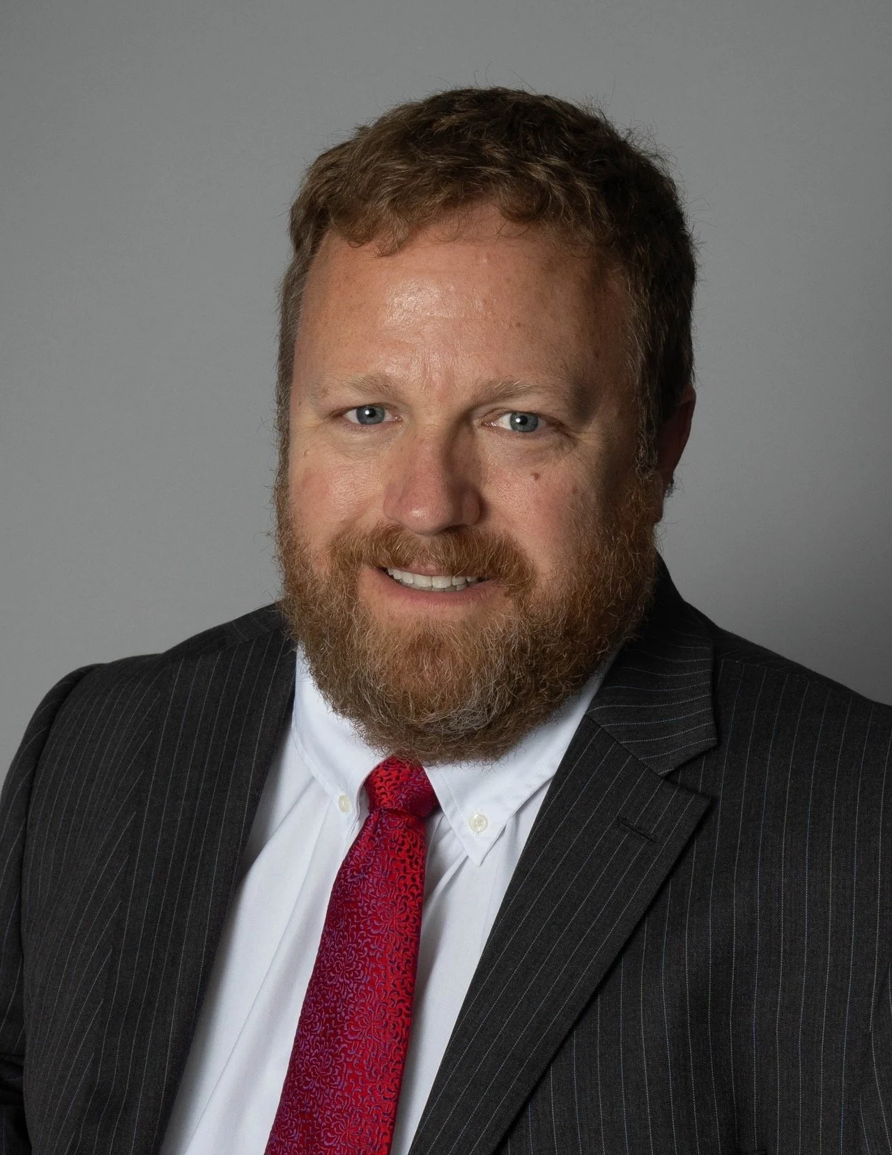 A professional headshot of a man with a beard, wearing a dark pinstripe suit, white shirt, and red patterned tie, against a plain gray background.