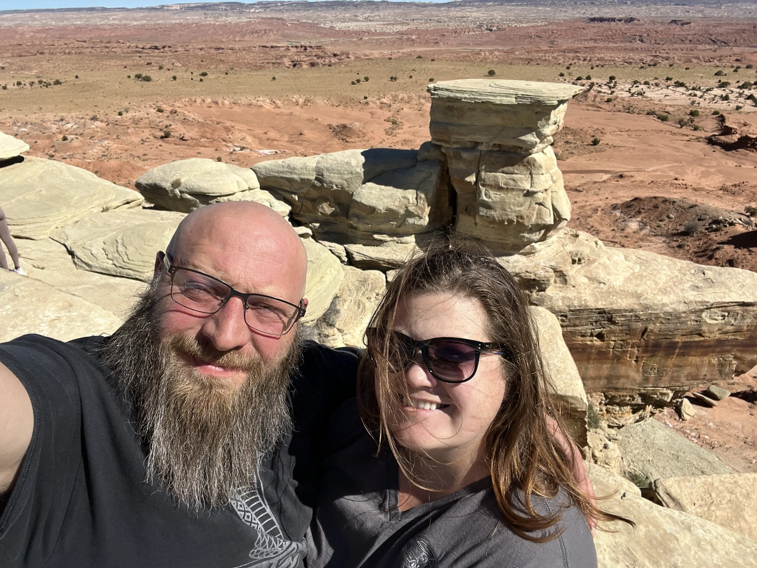 A man and woman taking a selfie in a desert landscape with large rocks and red rock formations in the background.