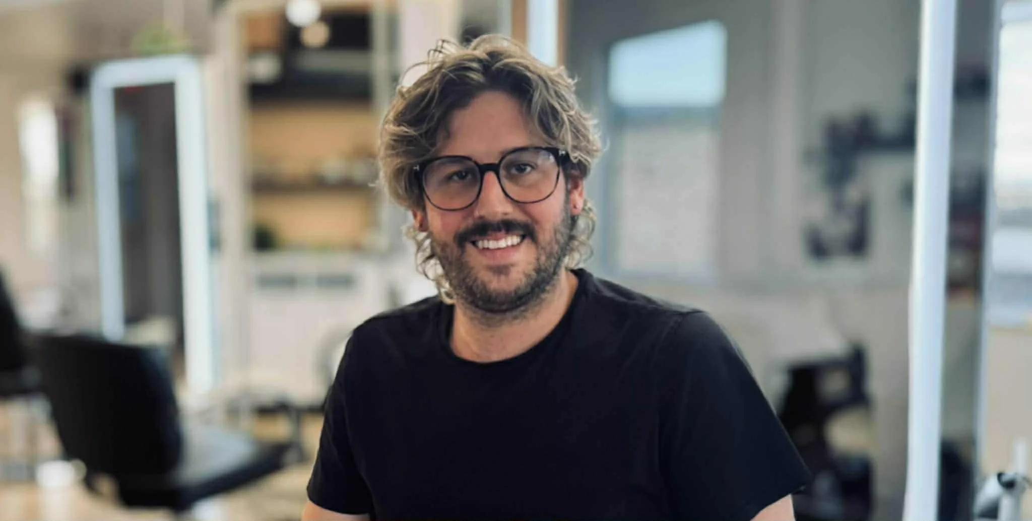 Un homme souriant avec des lunettes, cheveux bouclés et barbe, portant un t-shirt noir, dans un salon de coiffure et head spa de Longueuil.
