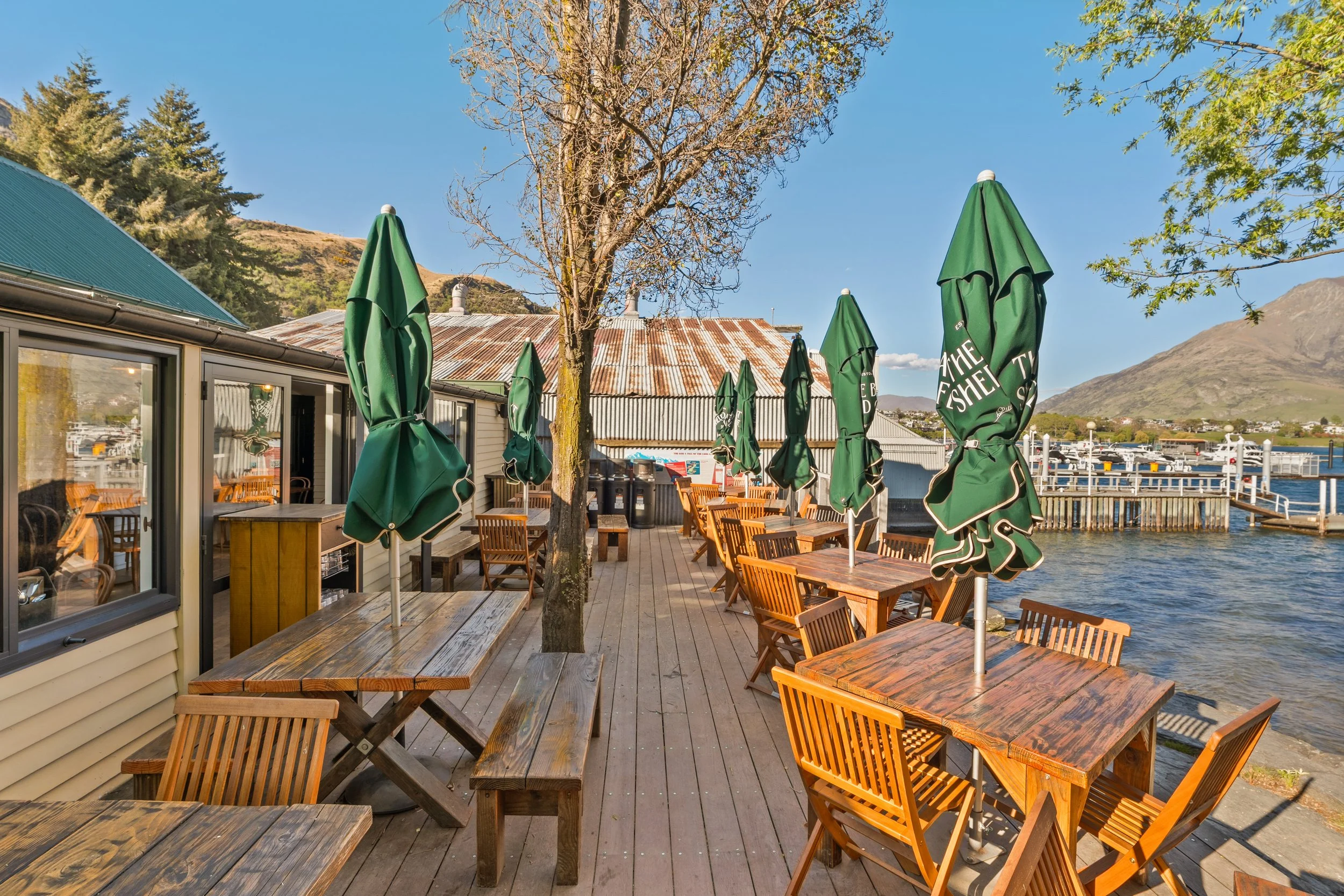 Lakeside deck with timber tables and green umbrellas looking to Lake Wakatipu and mountains