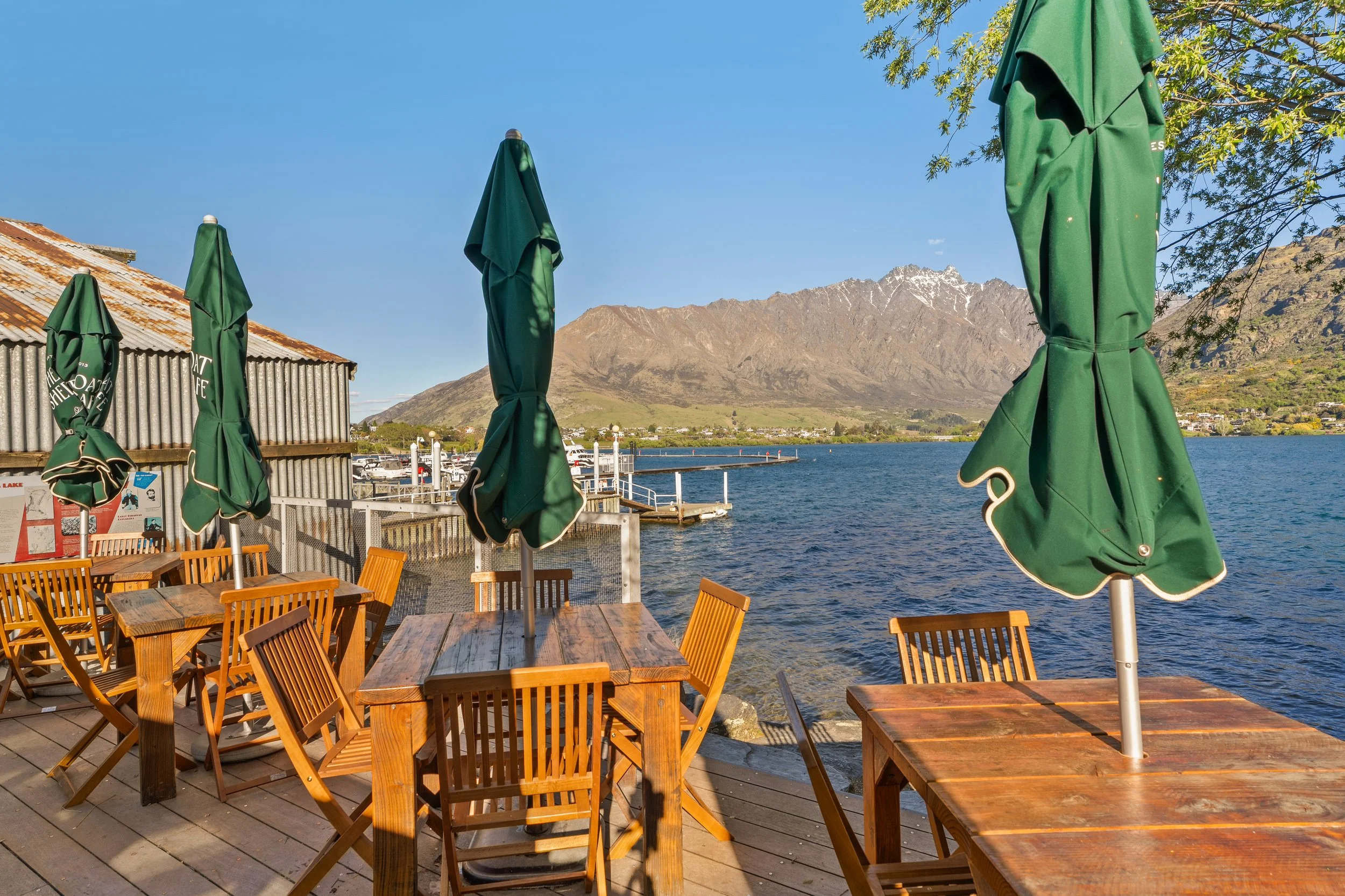 Lakeside deck with timber tables and green umbrellas looking to Lake Wakatipu and Remarkables mountain range