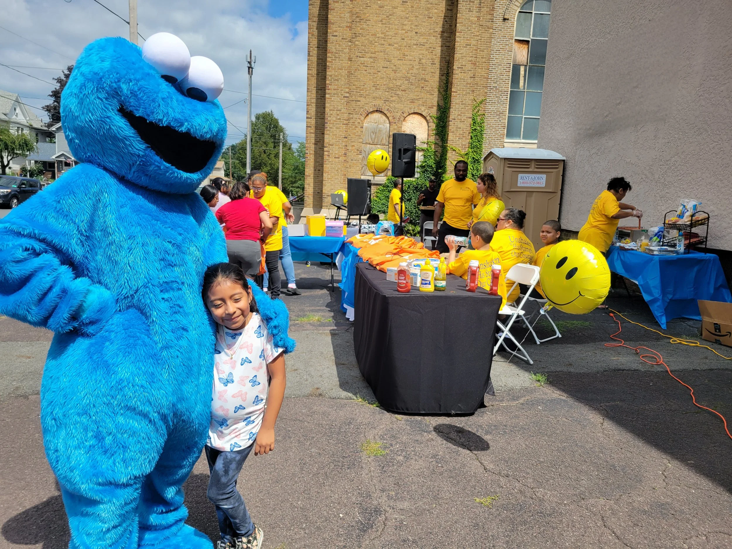 A child standing next to a person in a large blue Cookie Monster costume at an outdoor event. People are gathered around tables with food and balloons, with a brick building and cloudy sky in the background.