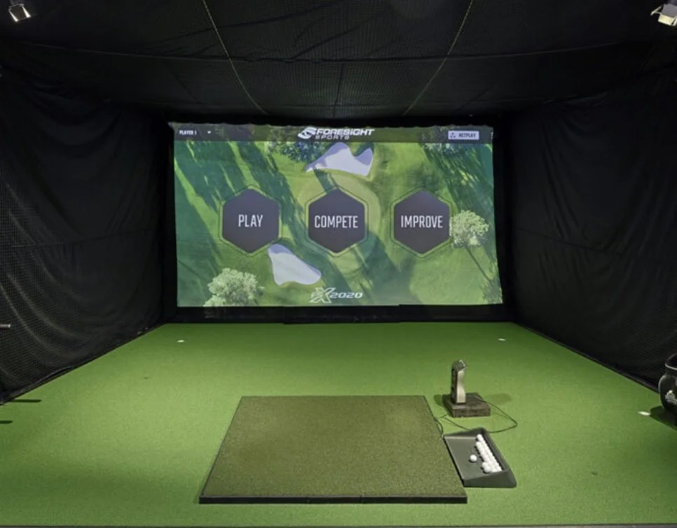 Indoor golf simulator room with a large screen displaying options to Play, Compete, and Improve, a putting green, golf clubs, and a golf ball collector.