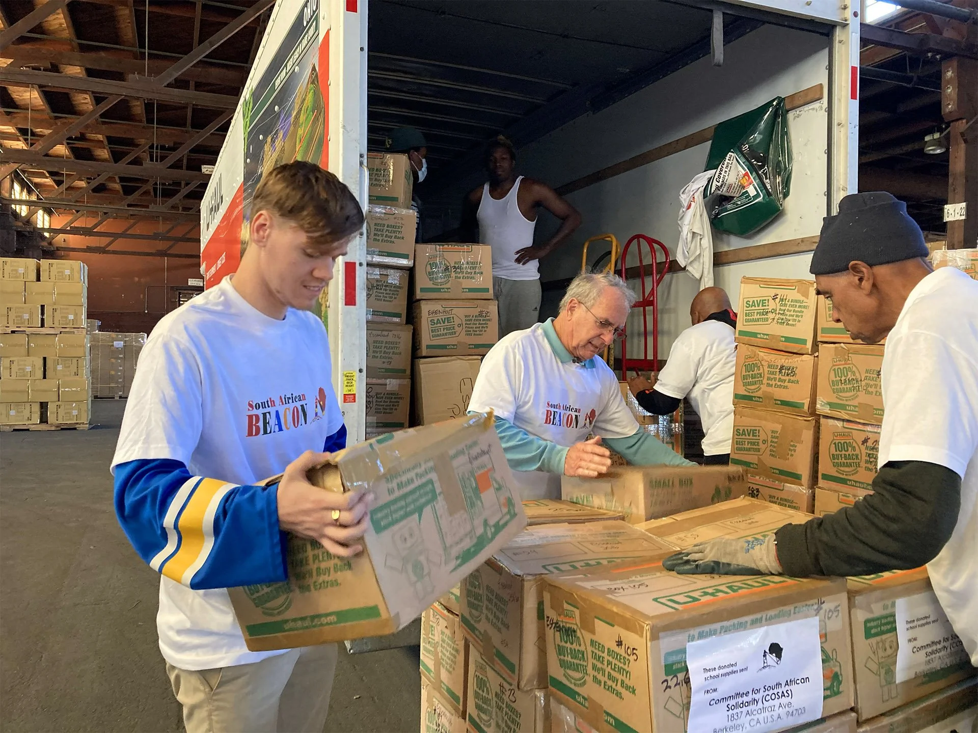 Volunteer Page Hero Image: Five guys, young and old, black and white, work as a team to load boxes of donated supplies in a large vehicle inside a warehouse.