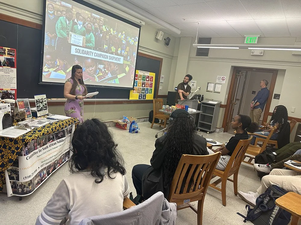 Photo of a COSAS organizer in front of a room speaking to what appear to be students sitting in wooden chairs. On the projector screen behind her is a photo of the COSAS "Solidarity Campaign Shipments"