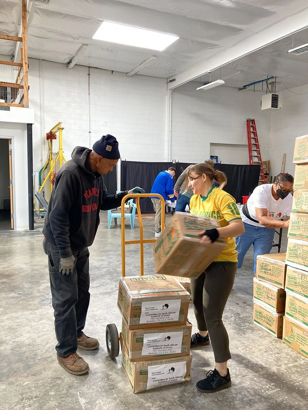A young woman loading boxes on a dolley while an older man wearing a blue beanie, wearing gloves, supervises.
