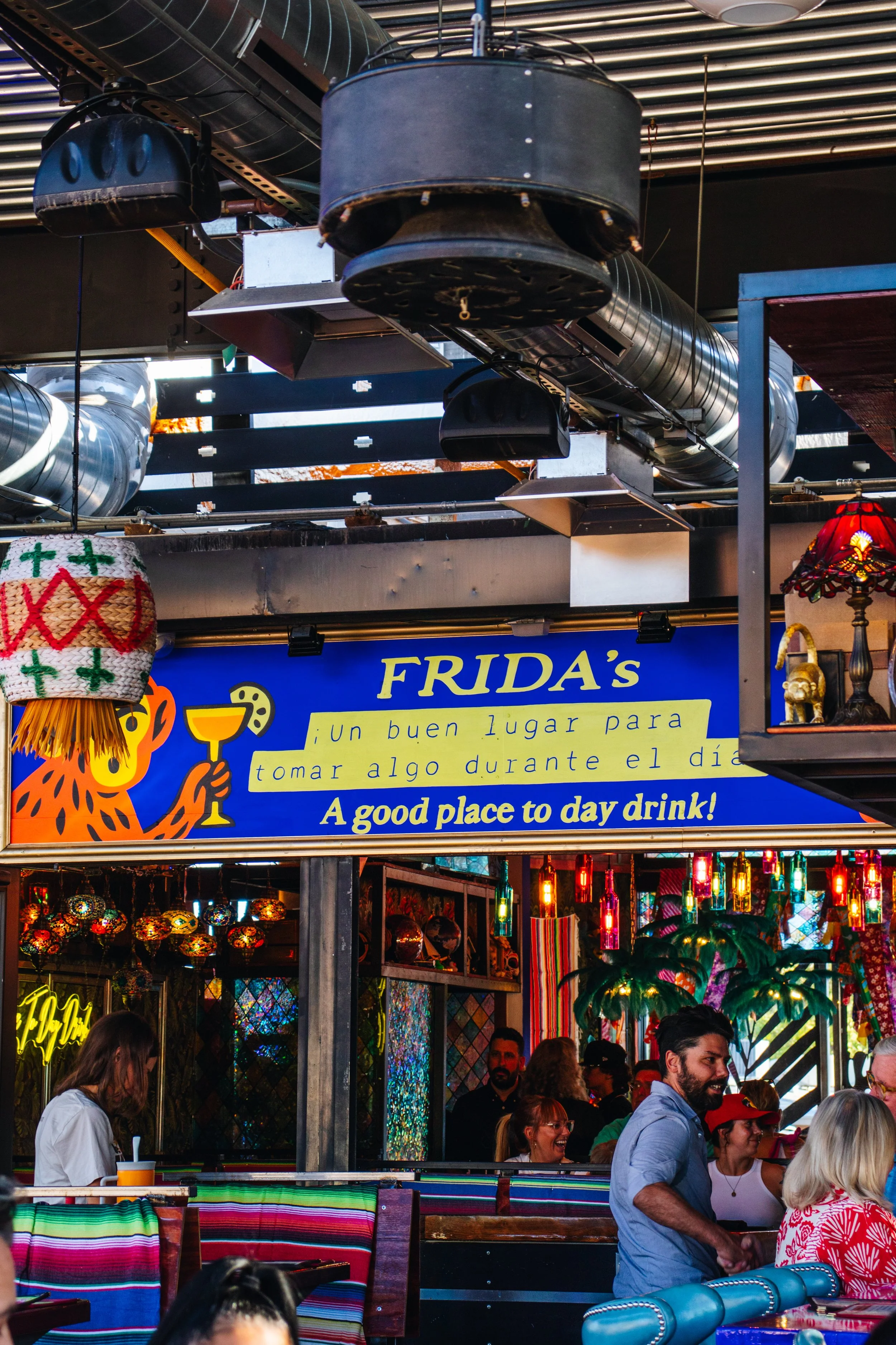 People gathered at an outdoor market stall with colorful Mexican decorations and lamps, with a blue sign that reads "FRIDA'S" and has text in Spanish promoting a good place for daytime drinking.