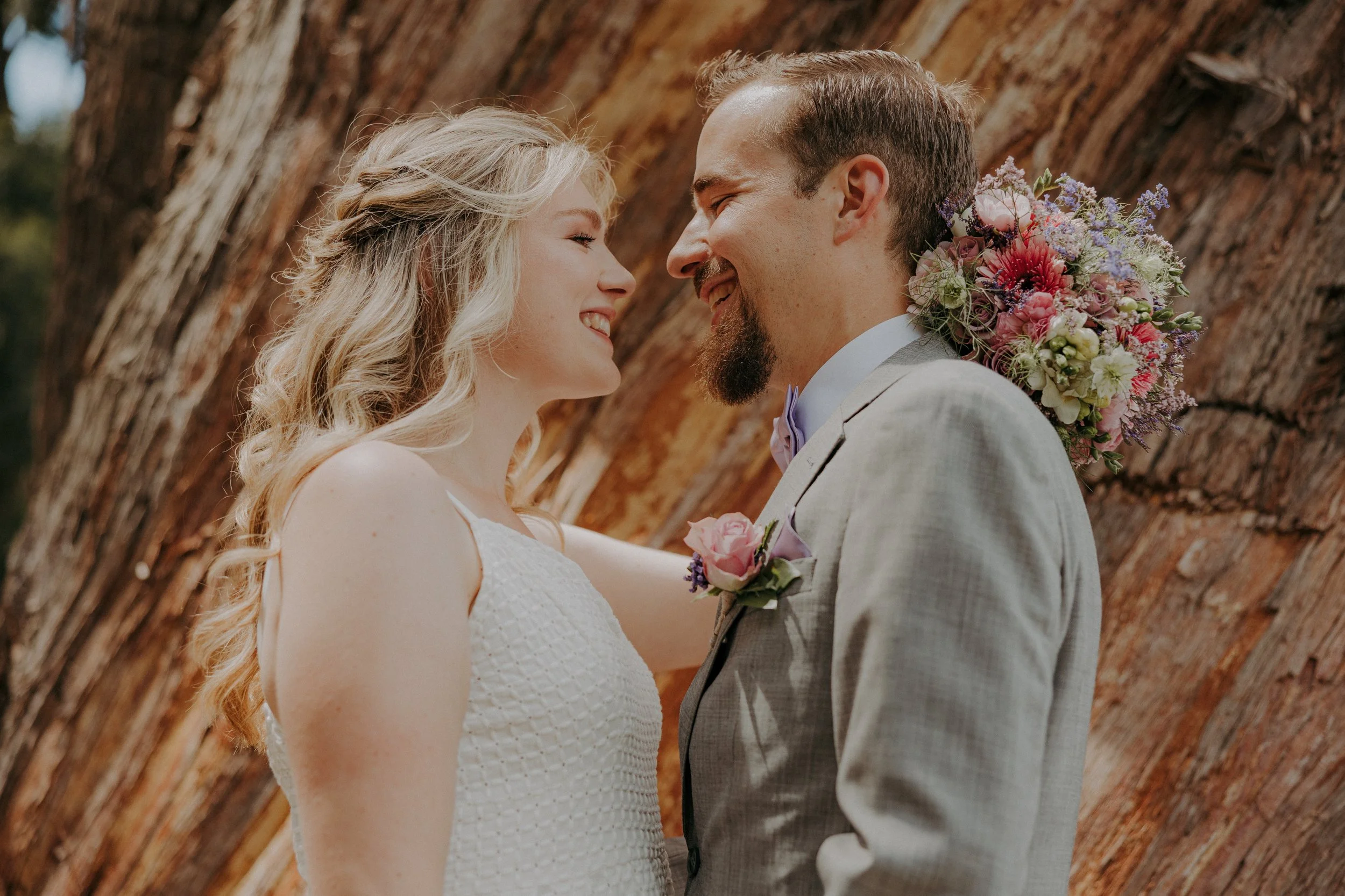 A couple on their wedding day, smiling and standing close with foreheads touching, in front of a large tree trunk.