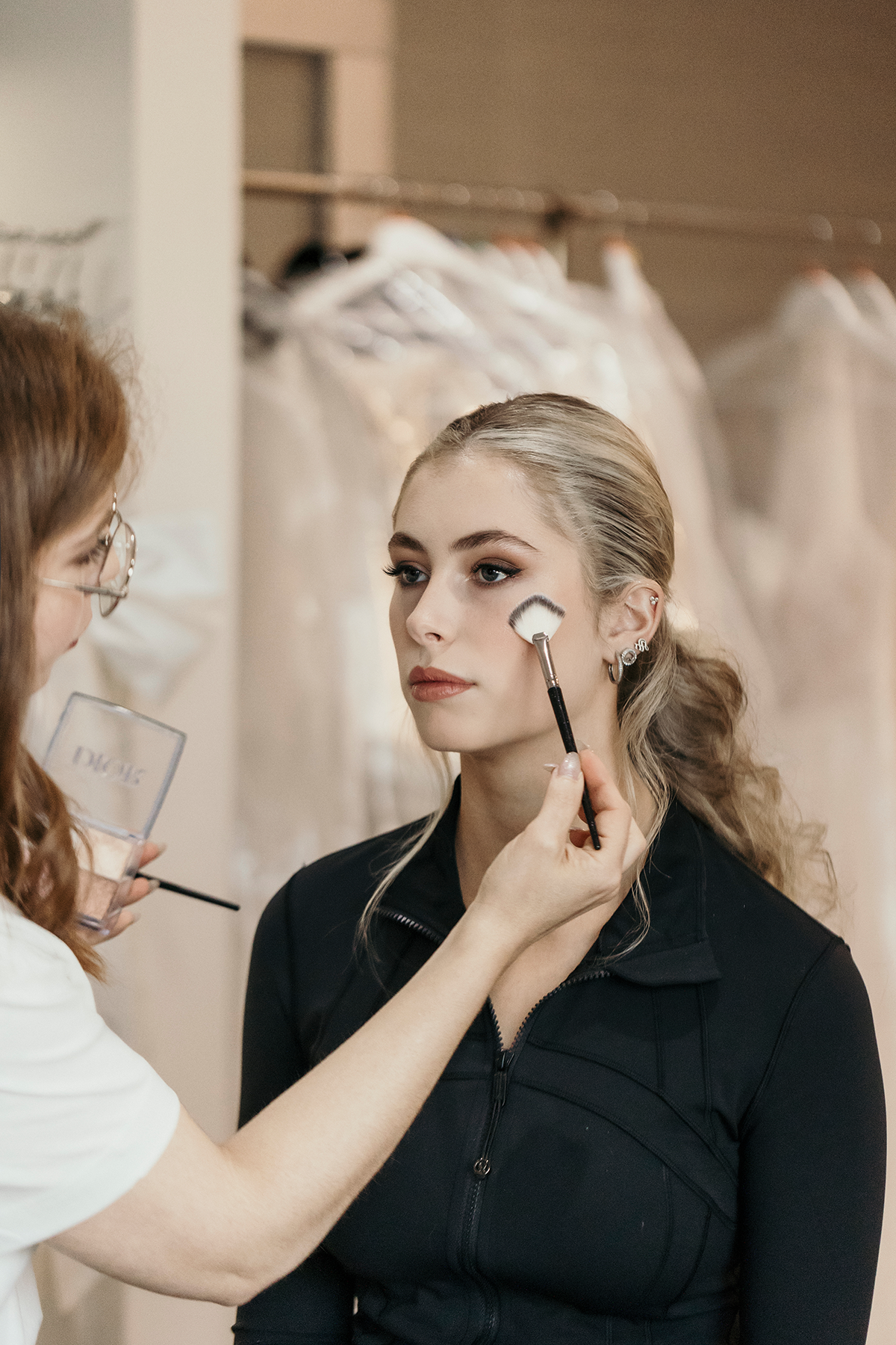 A woman with blonde hair getting makeup applied by a makeup artist in a dressing room with bridal gowns in the background.