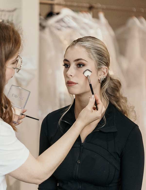 Soothing makeup application on a young woman with blonde hair in a ponytail, dressed in black, in a dressing room or boutique, with wedding dresses hanging in the background.