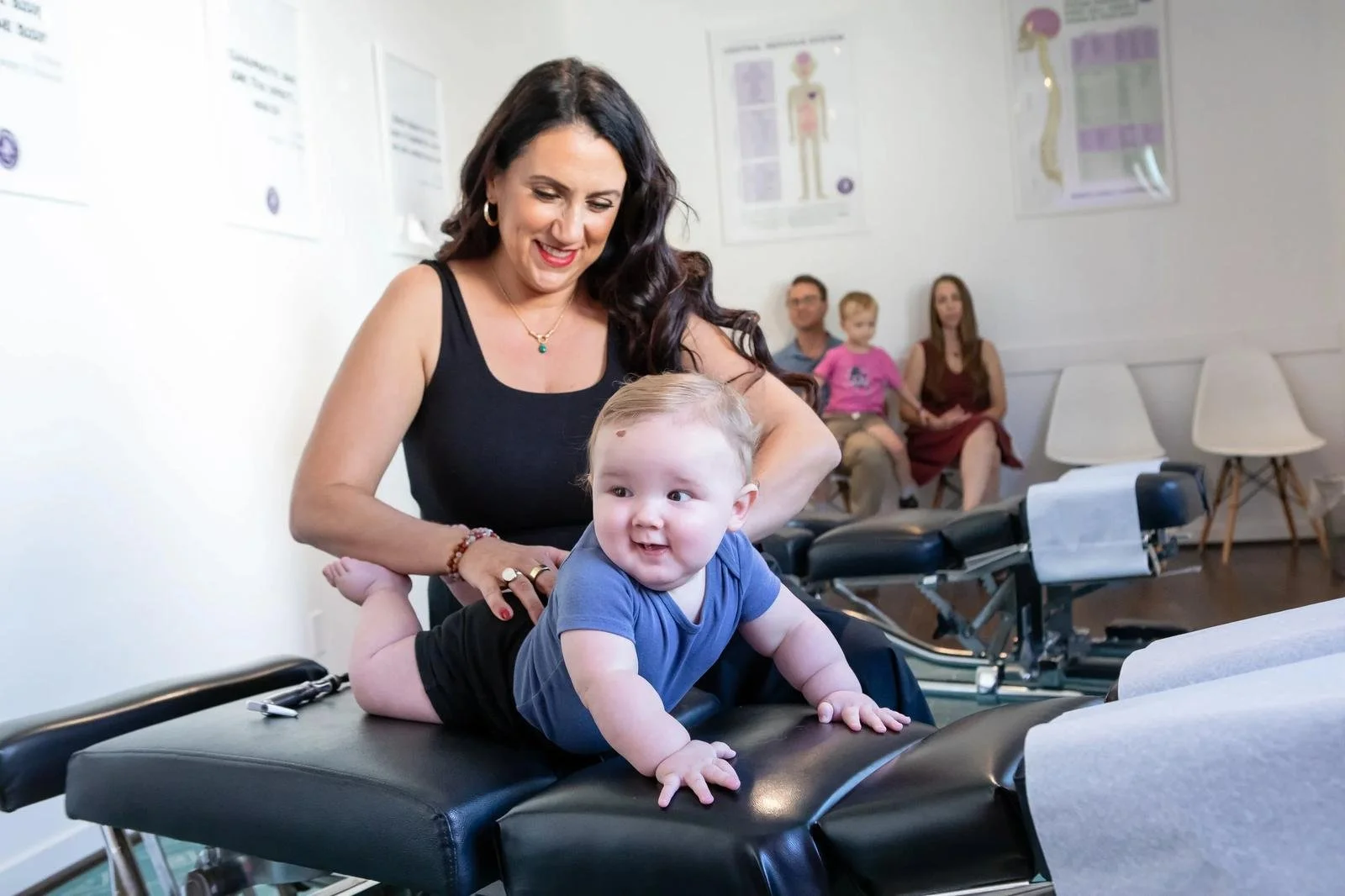A smiling baby on the chiropractic table at Innate Family Chiropractic Pasadena CA — Dr. Adana behind, family in background.