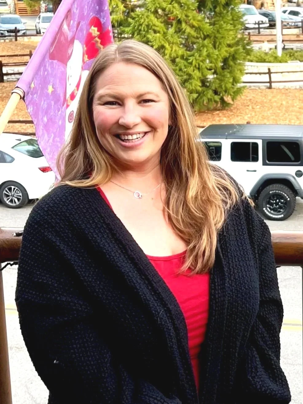 A woman with long blonde hair smiling outdoors. She is wearing a red top and a black jacket, standing in front of parked cars and a purple flag.