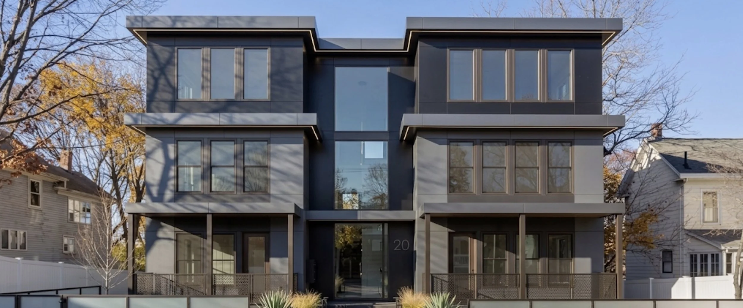 Modern black multi-story apartment building with large windows, situated in a neighborhood with trees and older houses, during autumn. DillCo Development Group