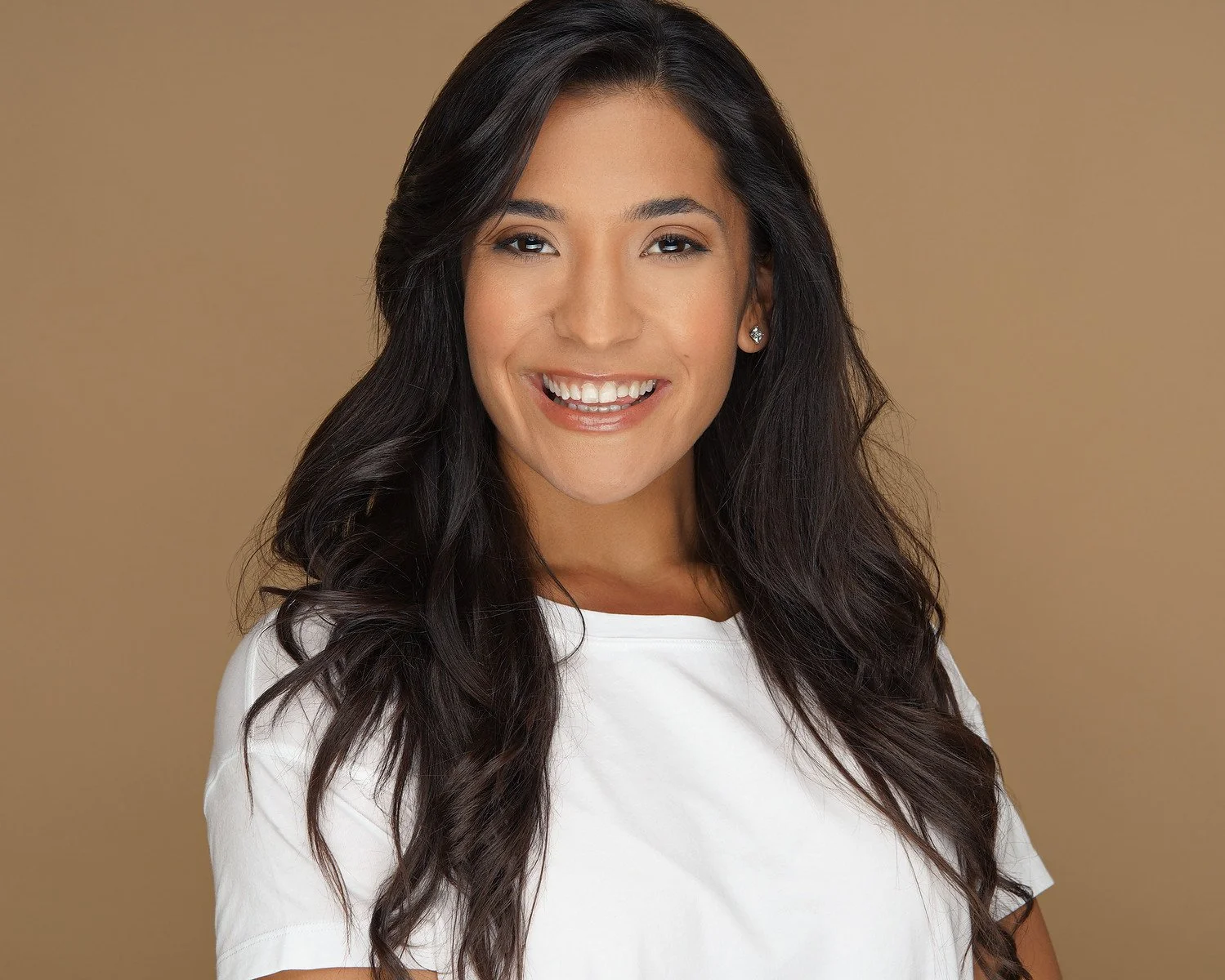 A woman with long, dark wavy hair smiling, wearing a white top and small earrings, against a plain beige background.