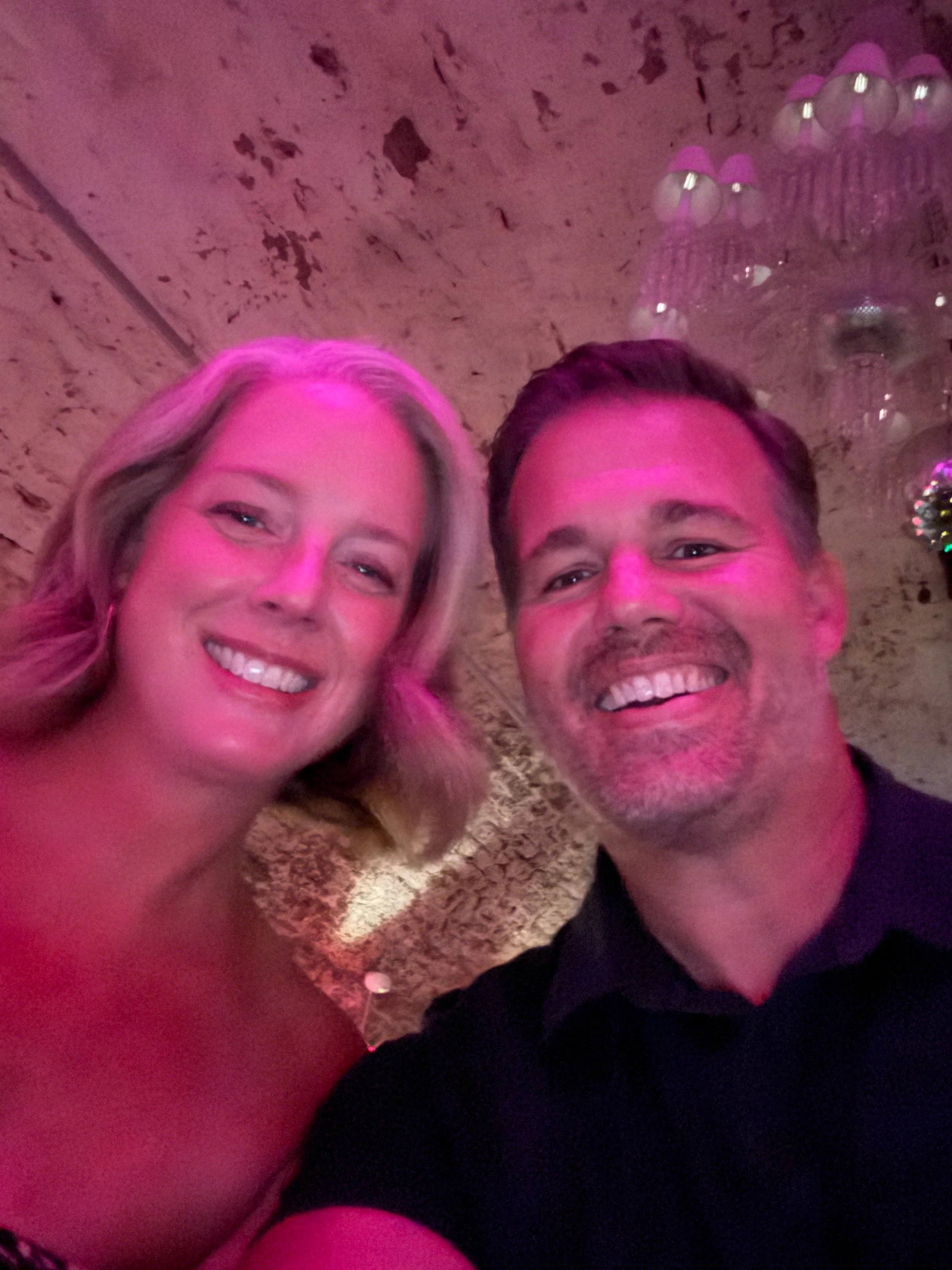 A smiling woman with blonde hair and a smiling man with short hair, both under pink lighting, taking a selfie in a decorated setting with a chandelier.