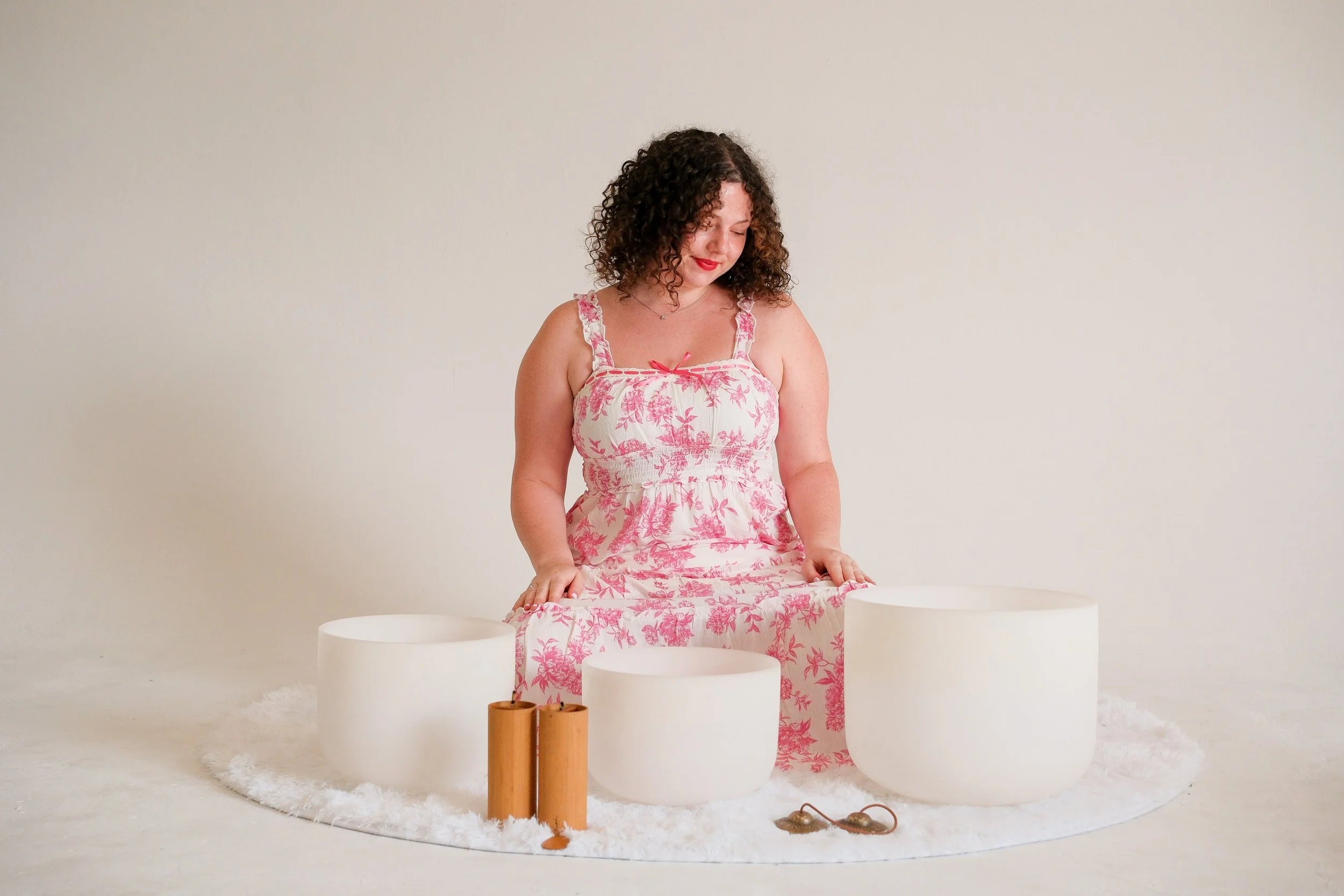 Woman in pink floral dress sitting on a fluffy white rug surrounded by white singing sound bowls and chimes and a with a white background.