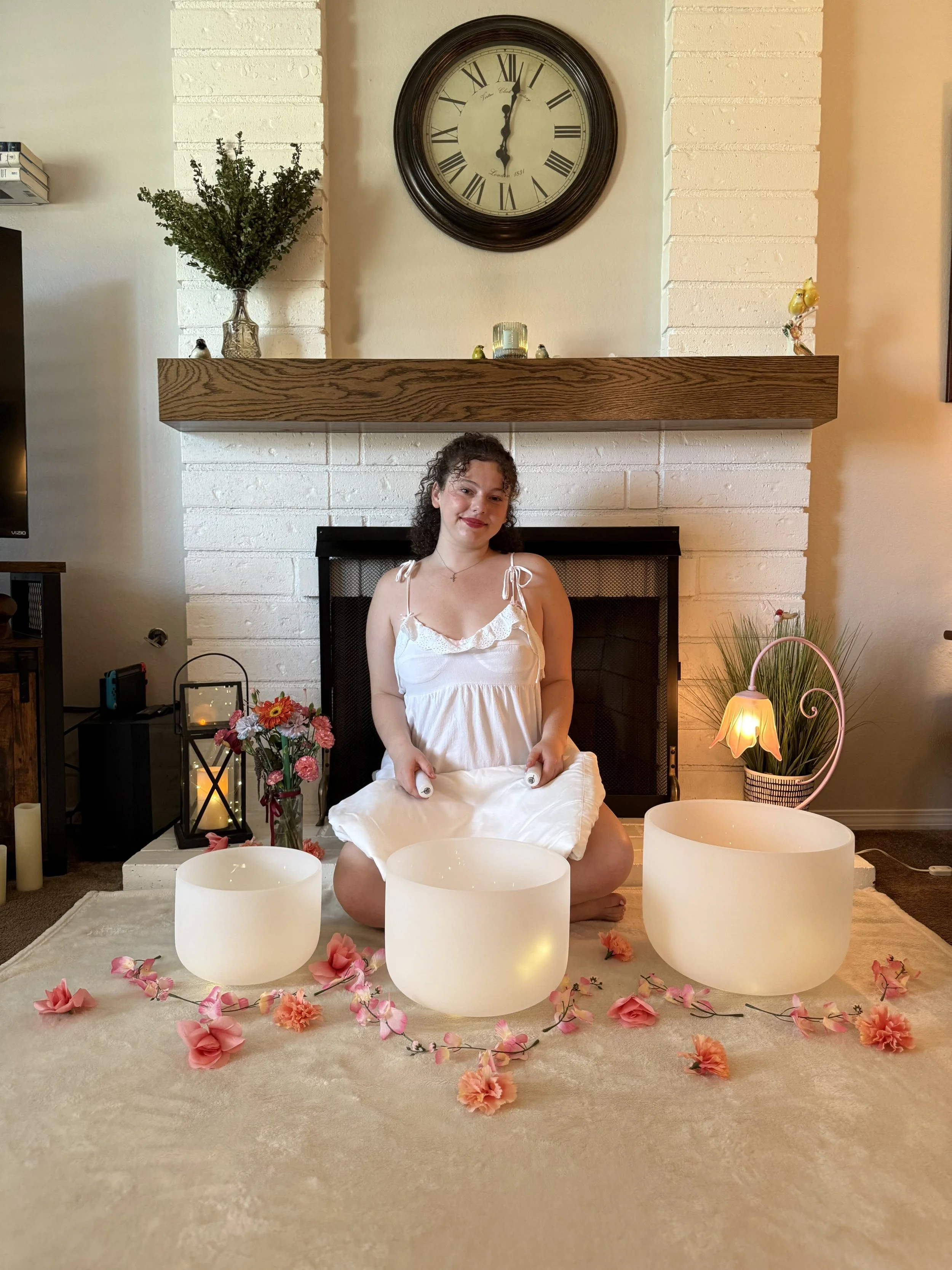 A woman kneeling on a beige carpet, holding white crystal singing bowls. She is surrounded by pink and peach flower garlands and loose flowers. Behind her is a white brick fireplace with a wooden mantel, decorated with candles, a vase with flowers, and small decorative objects. Floating candles and a potted plant are also part of the setting, with a large clock on the wall above the fireplace.