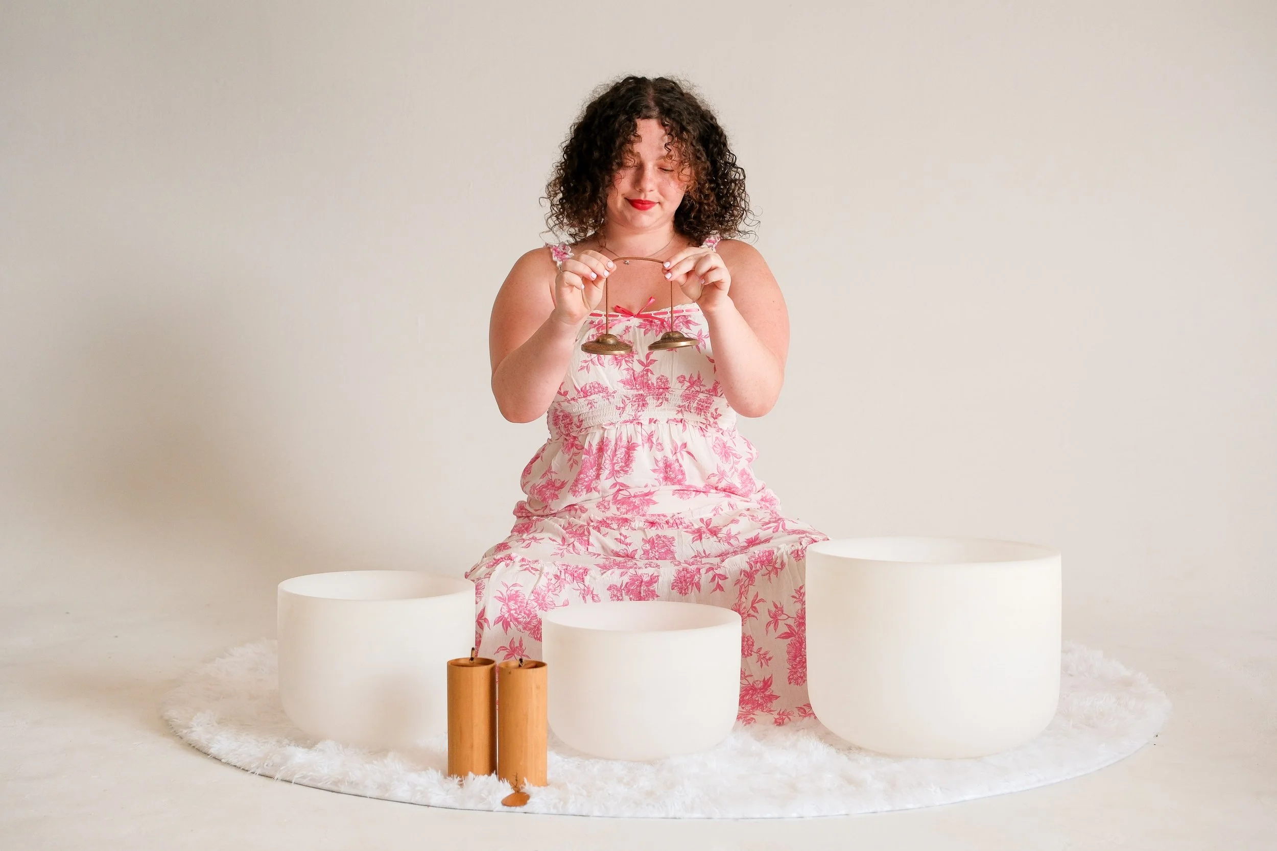 A woman with curly hair wearing a pink and white floral dress, sitting on a white fluffy rug, holding a tingsha, surrounded by white sound bowls and two wooden percussion chimes.