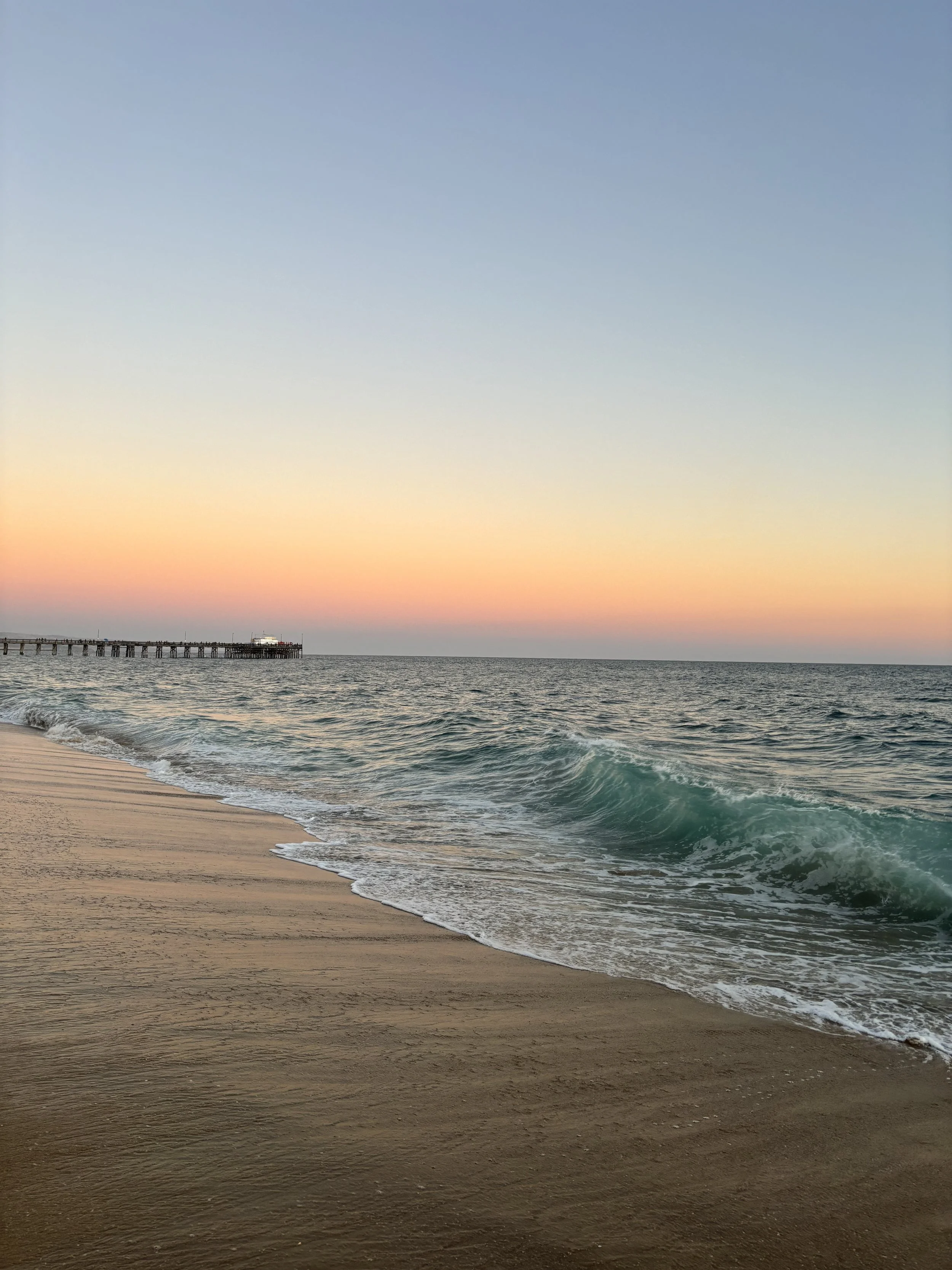 A sunset over the ocean with waves crashing on a sandy beach and a pier extending into the water.