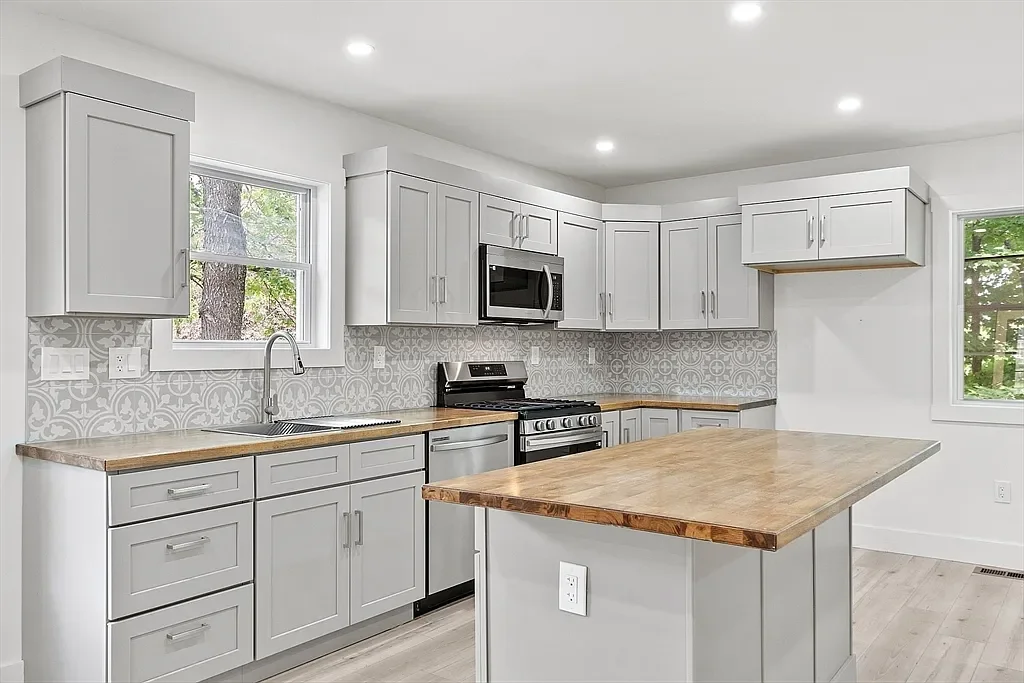 Modern kitchen with white cabinets, a wooden countertop island, stainless steel appliances, a decorative backsplash, and two windows showing greenery outside.