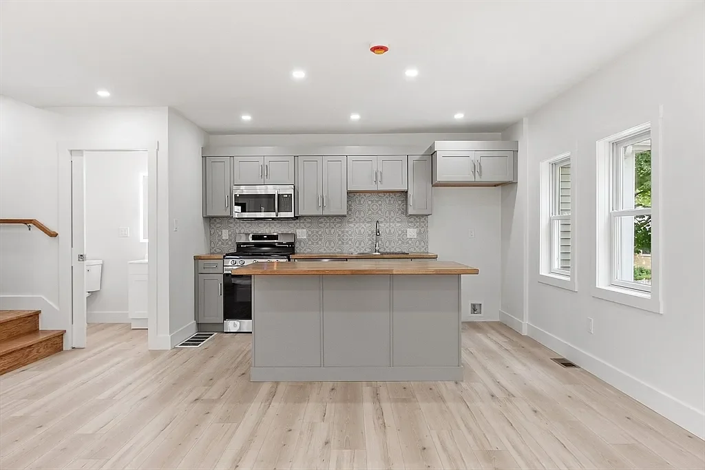 Modern kitchen with grey cabinets, a wooden countertop island, stainless steel appliances, and white walls with natural light from multiple windows.