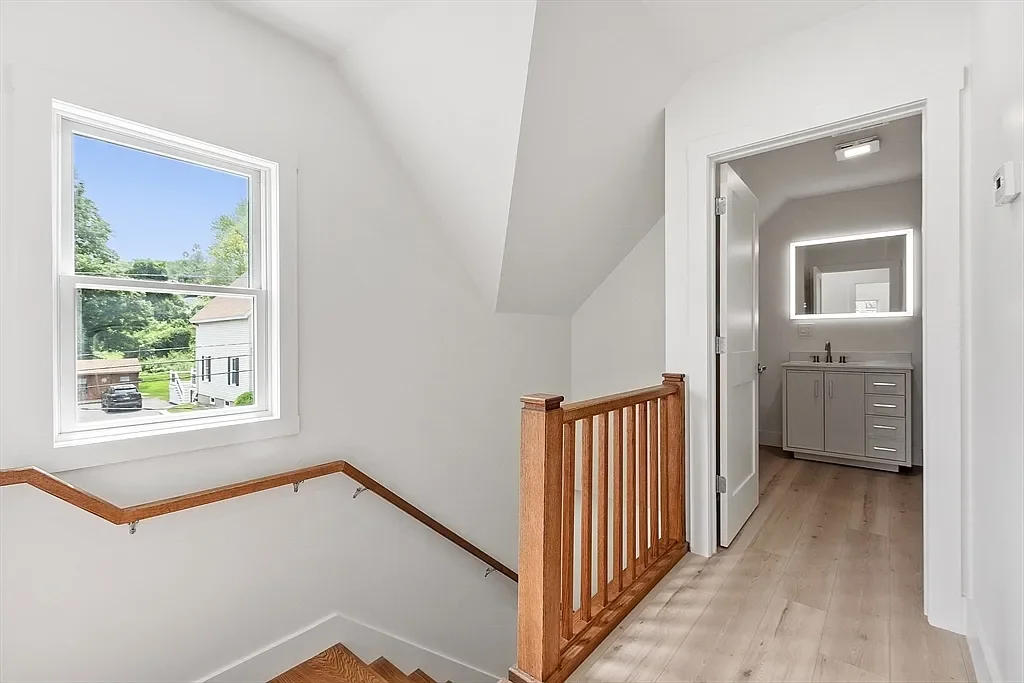 Upper hallway with a staircase, wooden handrail, window showing greenery outside, and a bathroom with a backlit mirror.