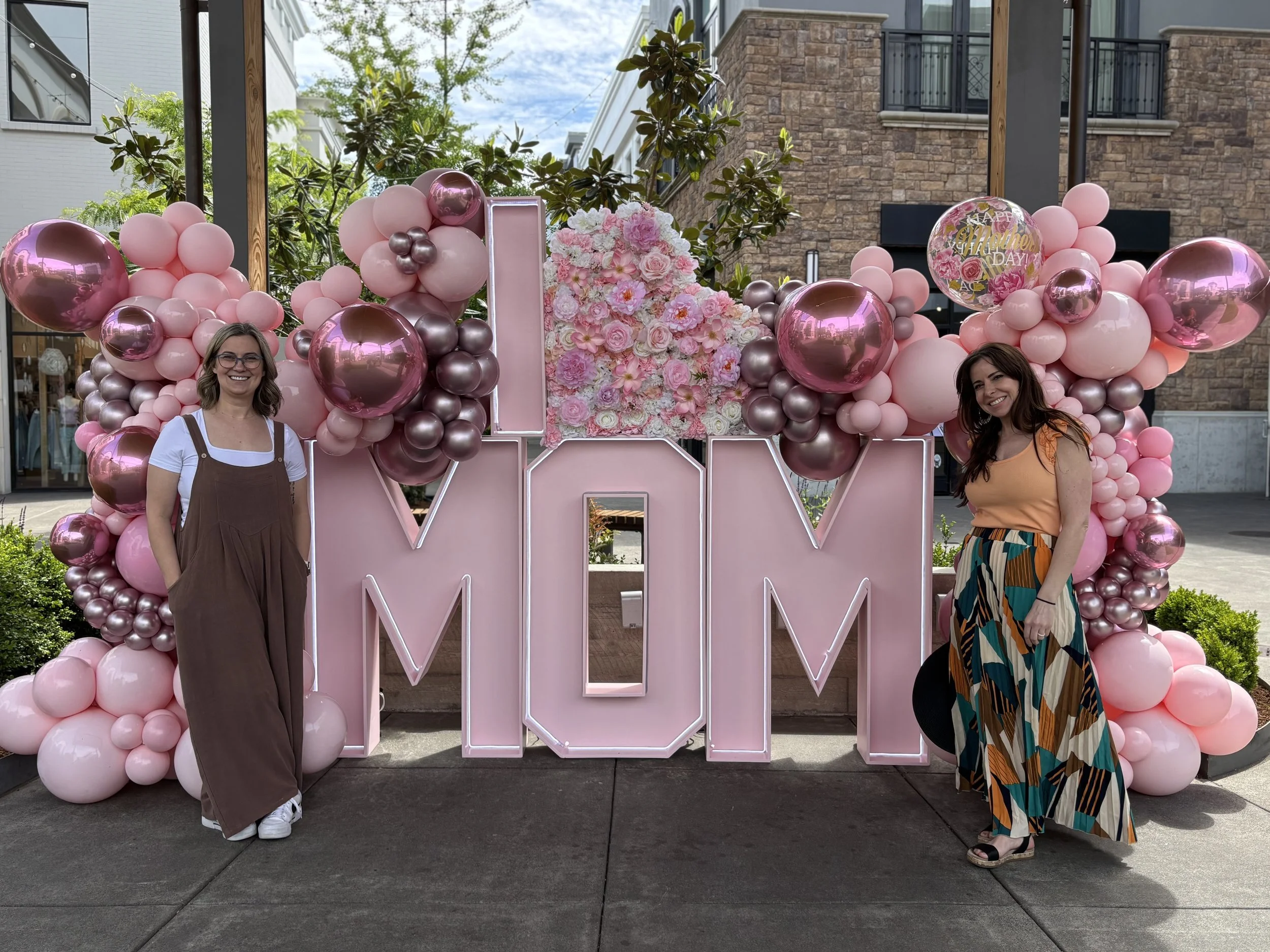 Two women standing on either side of a pink and floral balloon display that spells out 'MOM'. One woman is wearing a brown jumpsuit with a white shirt underneath, and the other is wearing a colorful skirt and orange top. The display features pink, purple, and metallic balloons, and a floral heart shape on top. There are buildings and trees in the background.
