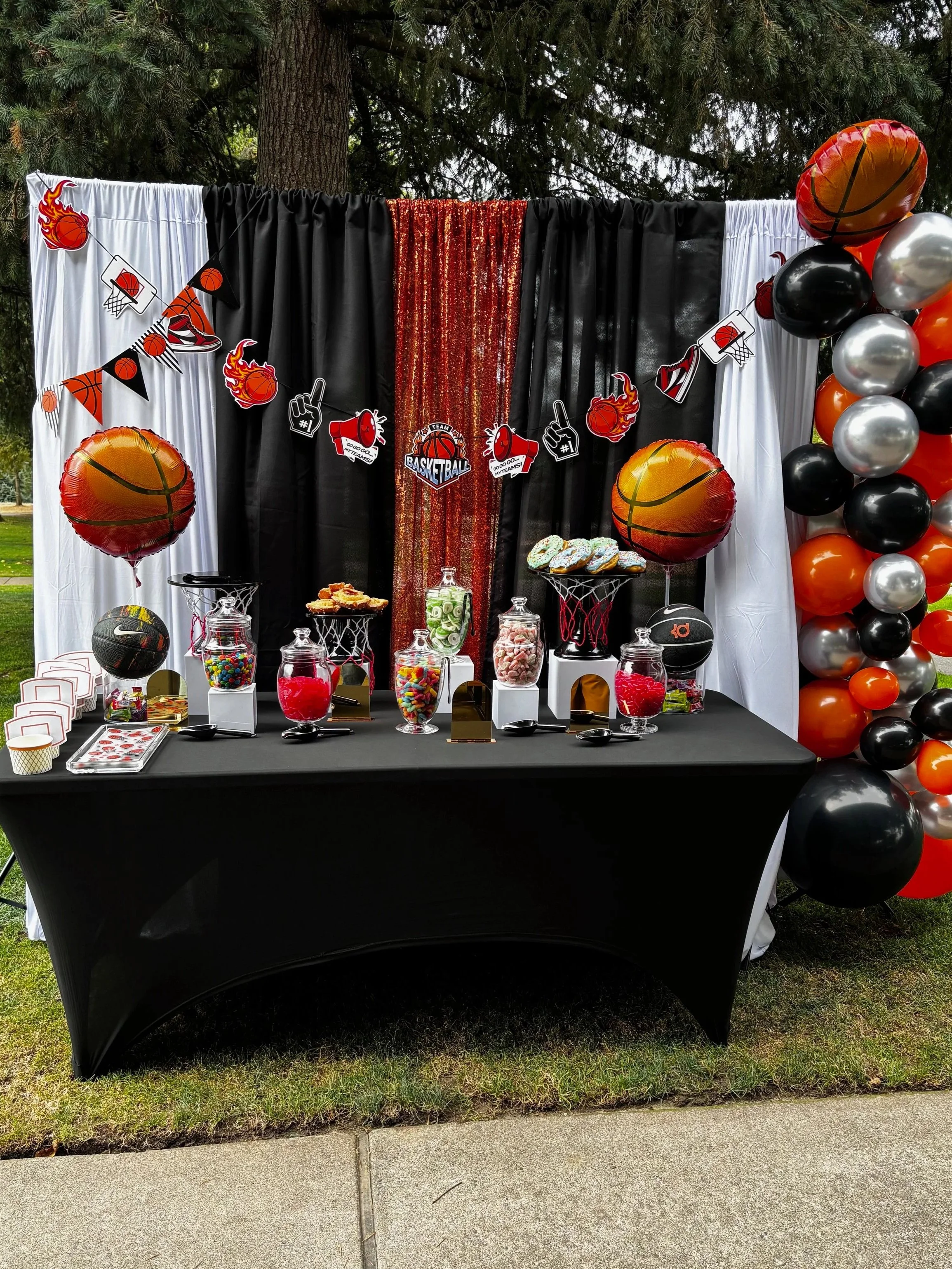 Basketball-themed party table with red, black, and white decorations, balloons, candy jars, and basketball-themed banners.