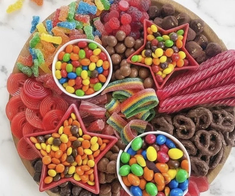 Colorful assortment of candies on a round wooden platter, including gummy worms, chocolate balls, rainbow sour strips, and jellybeans in star and bowl shapes.