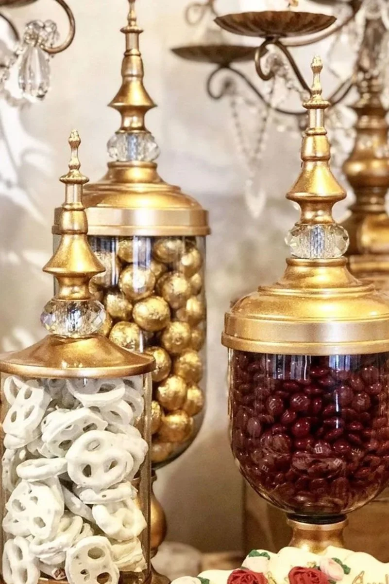 Decorative jars filled with white pretzels, gold foil balls, and red beans, topped with ornate gold and crystal finials.
