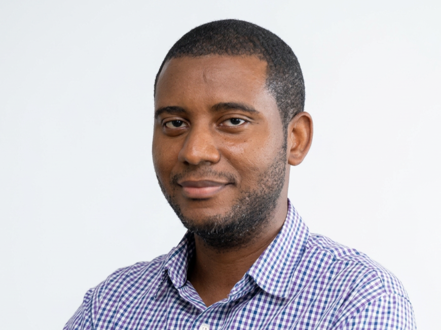 A man with short black hair and a beard, wearing a purple and white checkered shirt, looking at the camera with a slight smile against a plain white background.