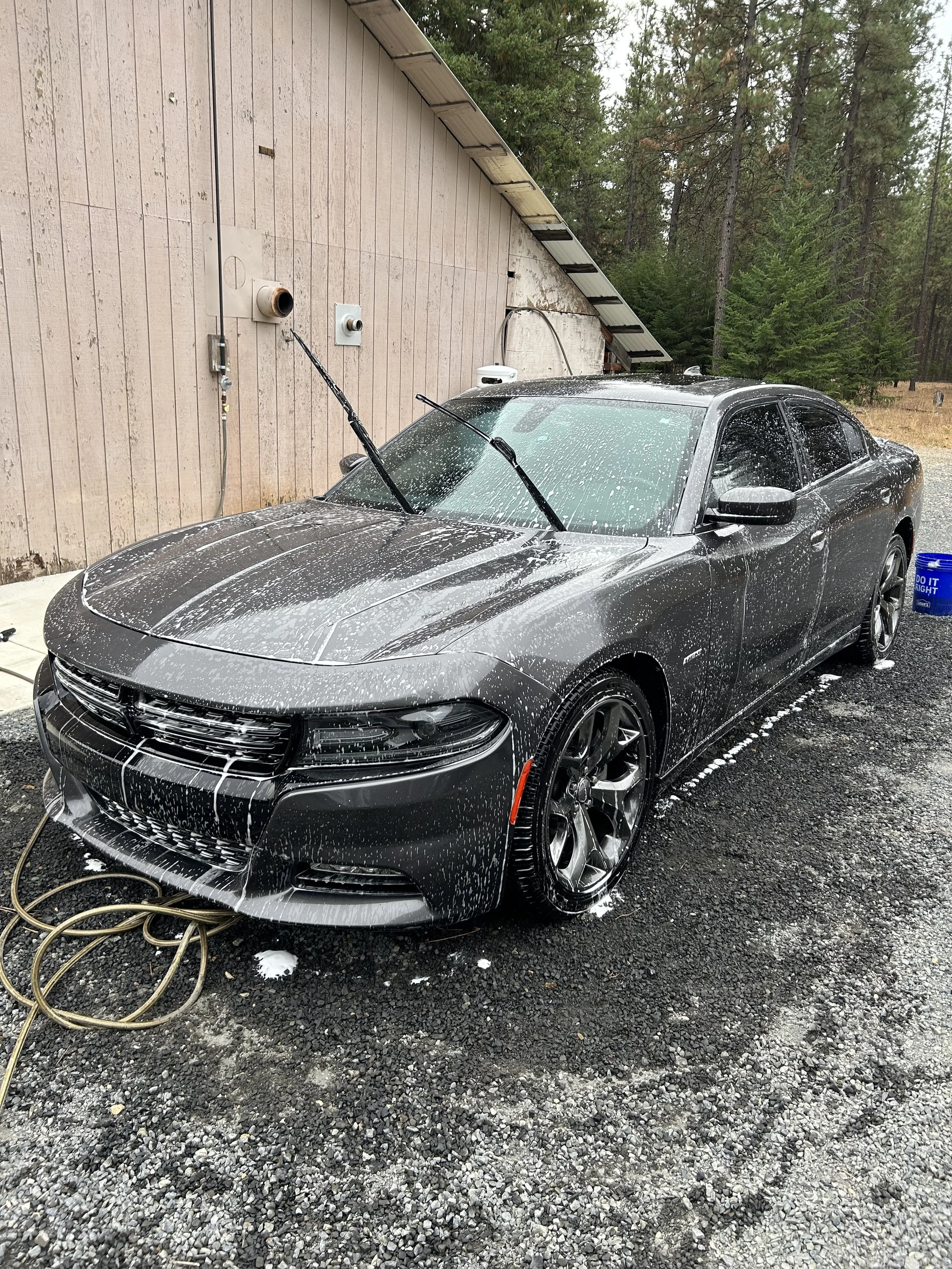 A black sedan car being washed with soap and water outside near a pink wooden building with trees in the background.