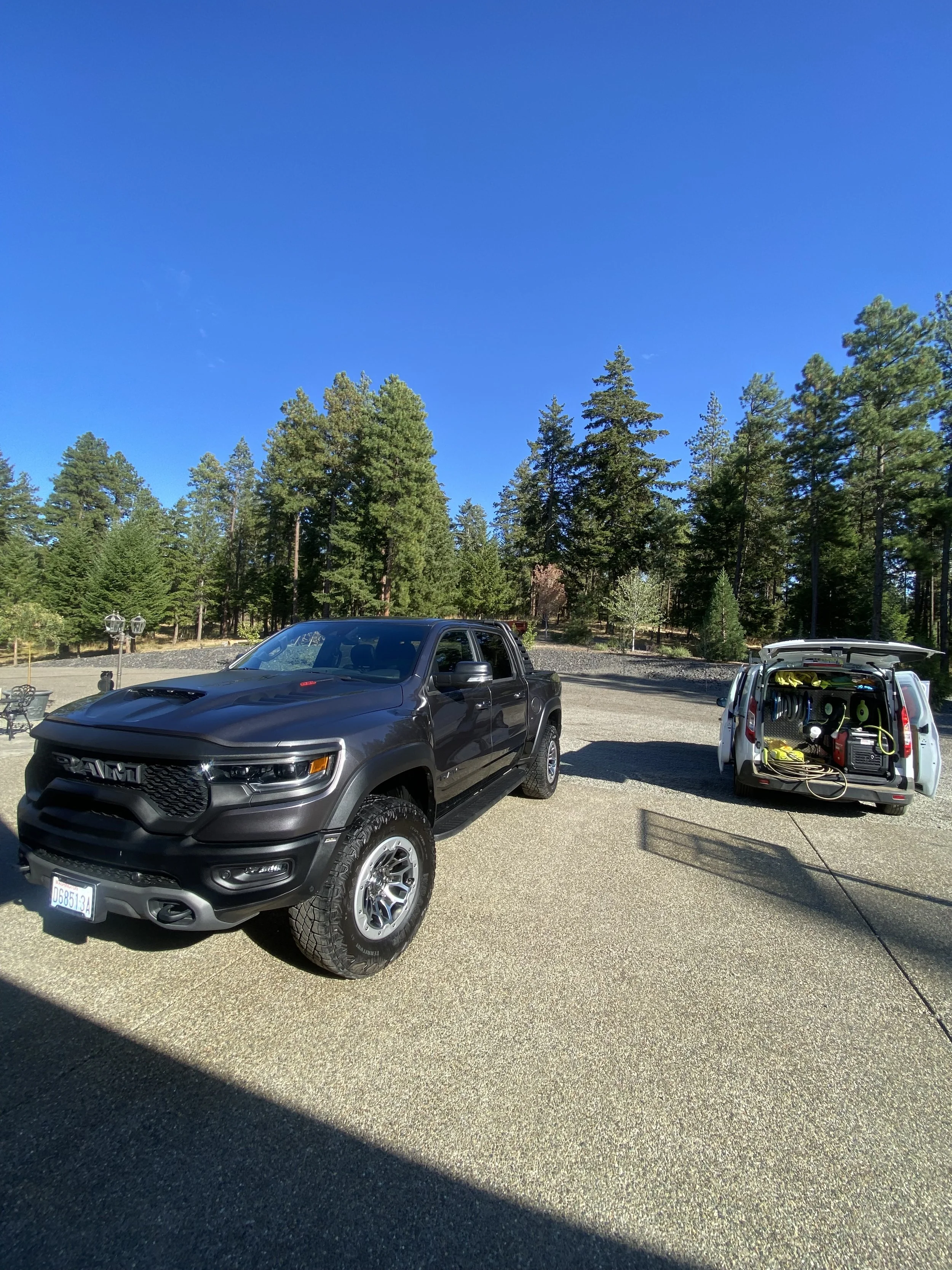 A black pickup truck parked next to a white work van with open doors revealing tools and equipment, with a forest of pine trees and a clear blue sky in the background.