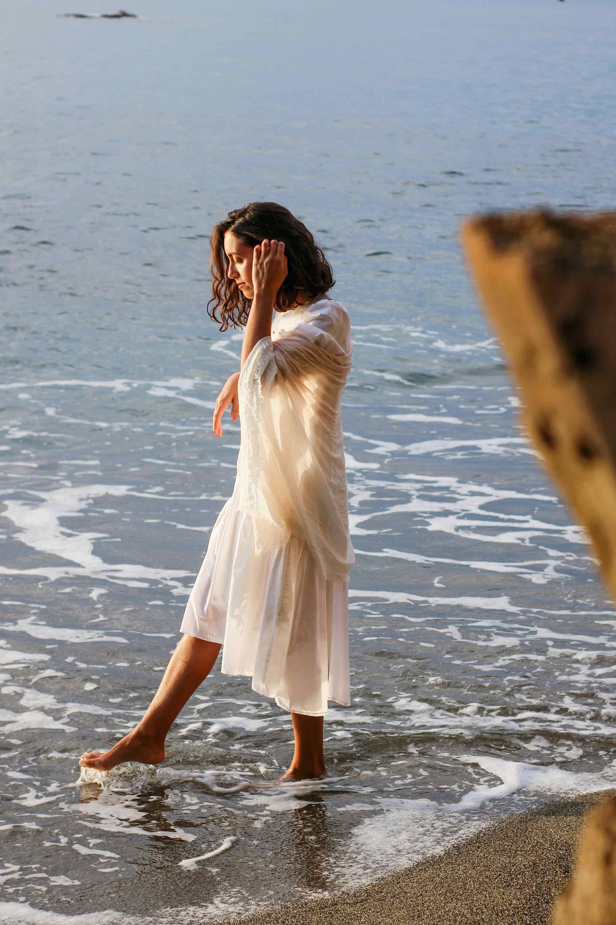 A woman in a light-colored dress with long sleeves, walking barefoot in the shallow water at the beach during sunset or sunrise.
