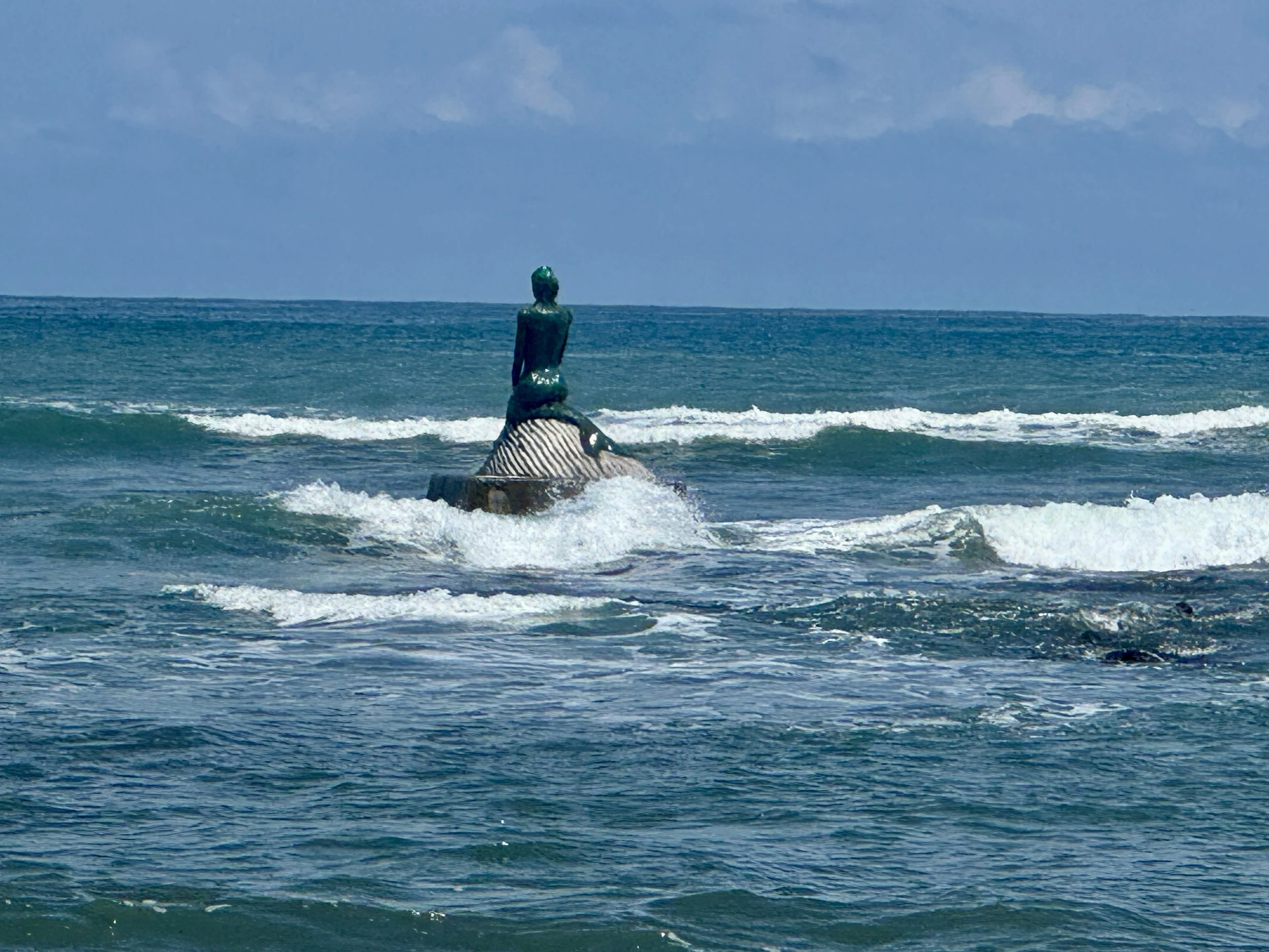 Mermaid statue perched on a rock in the ocean at La Sirena de Esterillos, surrounded by rolling waves.