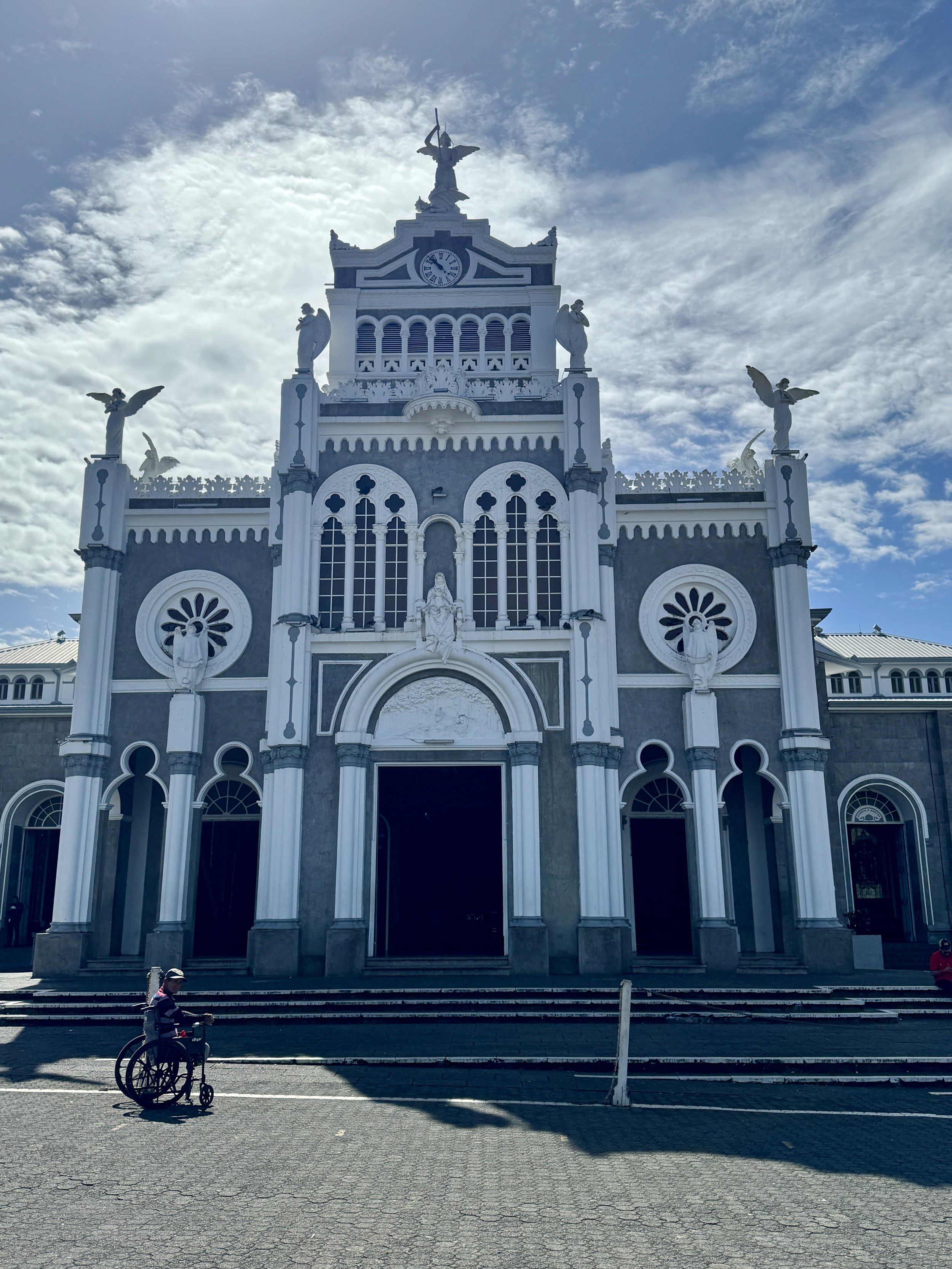 Front exterior of Basílica de Nuestra Señora de los Ángeles in Cartago with ornate white façade and statues against a blue sky.
