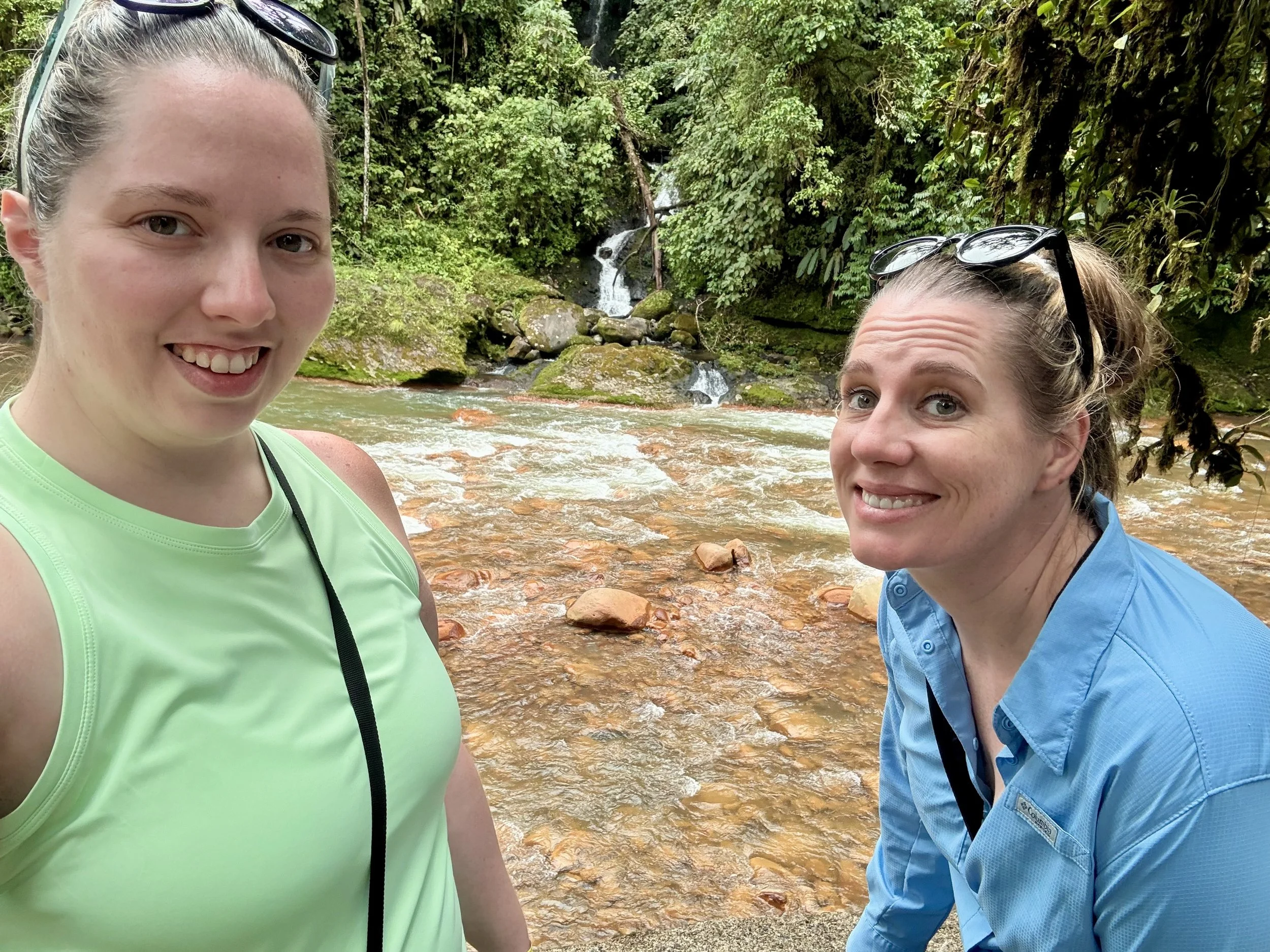 Two visitors smiling near a shallow river on the Recreo Verde property.