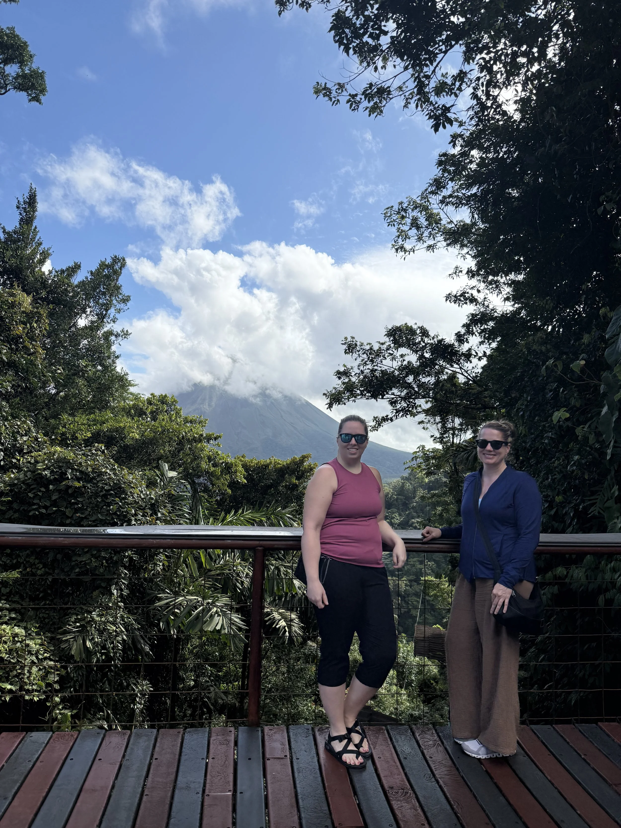 Two visitors posing at a scenic overlook with Lake Arenal and clouds behind them.